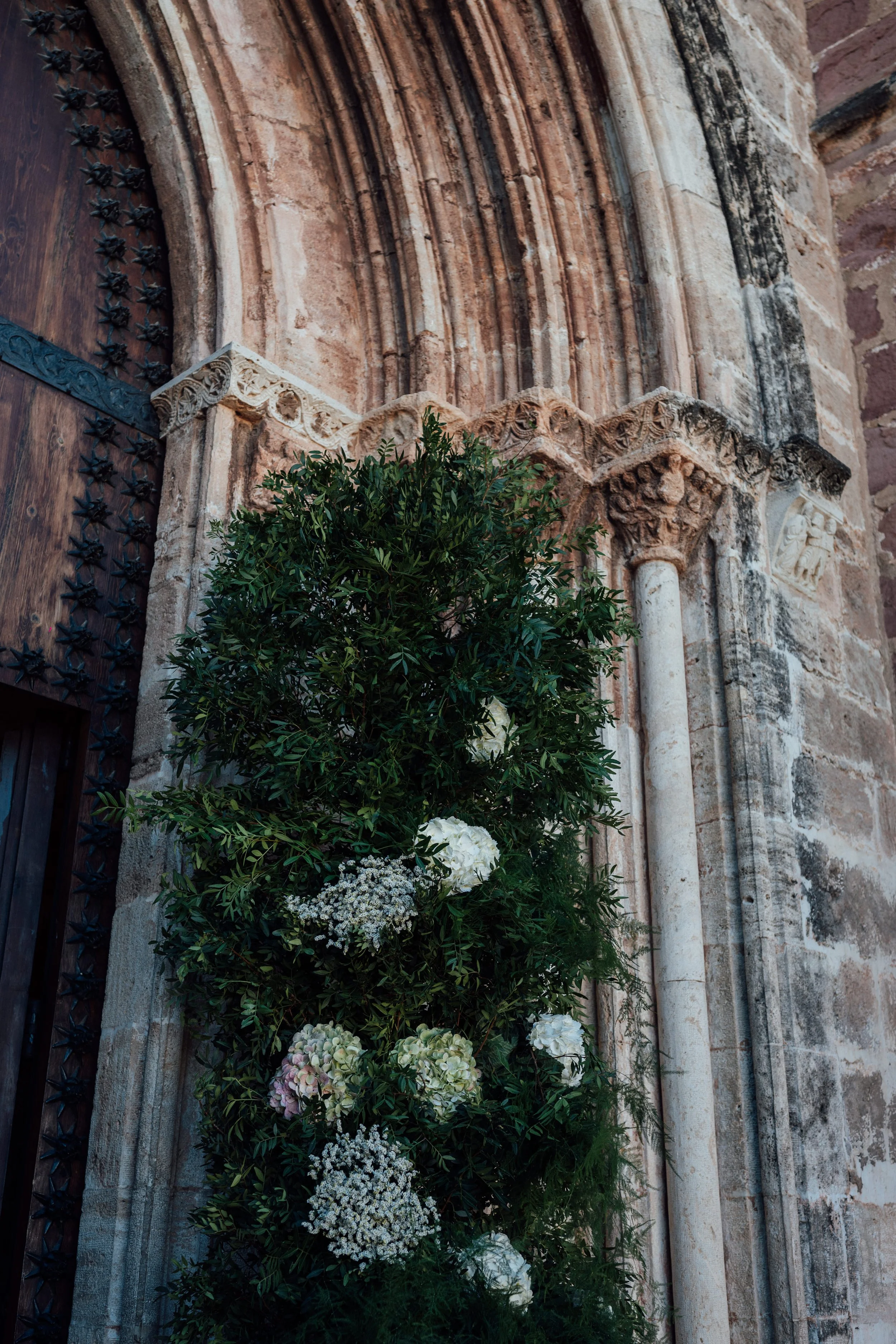 Detalles de una fachada antigua de piedra con columnas y arcos, adornada con flores y plantas verdes.