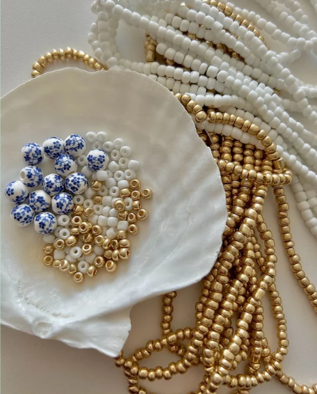 Various white, gold, and blue beaded necklaces and bracelet strands arranged around a scallop shell with loose beads inside.