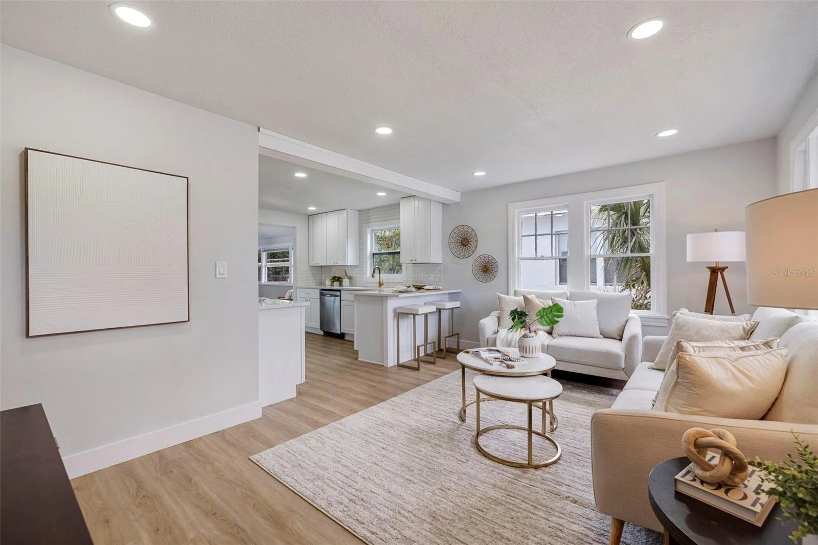 Living room with white sofas, beige pillows, round marble coffee tables, a tall floor lamp, and large windows, with a view into a kitchen with white cabinets and stainless steel appliances.