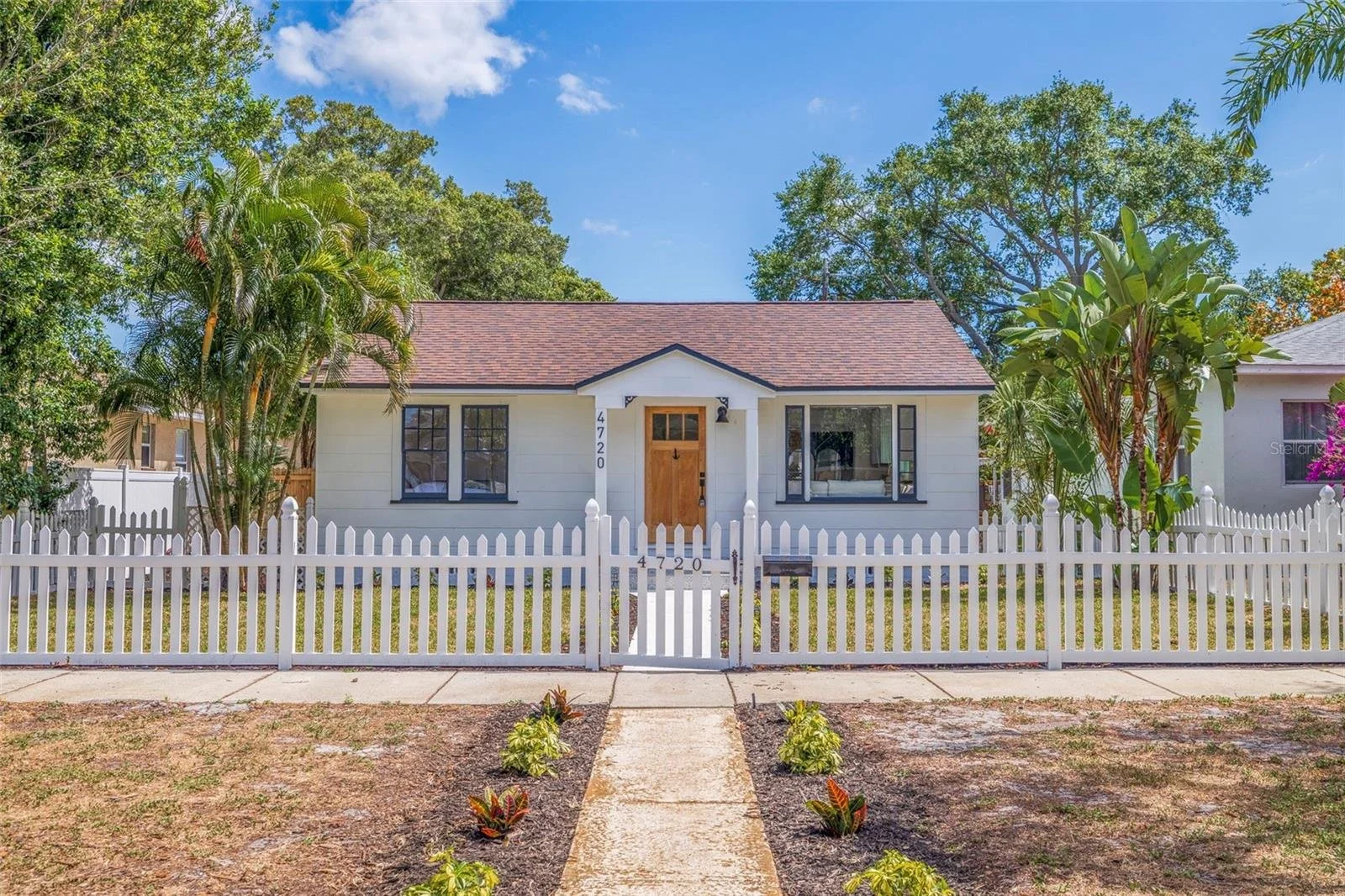 Front view of a white house with a brown front door, black window frames, and a small front porch. White picket fence surrounds the house. There are trees and bushes in the yard, with a sidewalk leading up to the entrance. The house number is 4720.