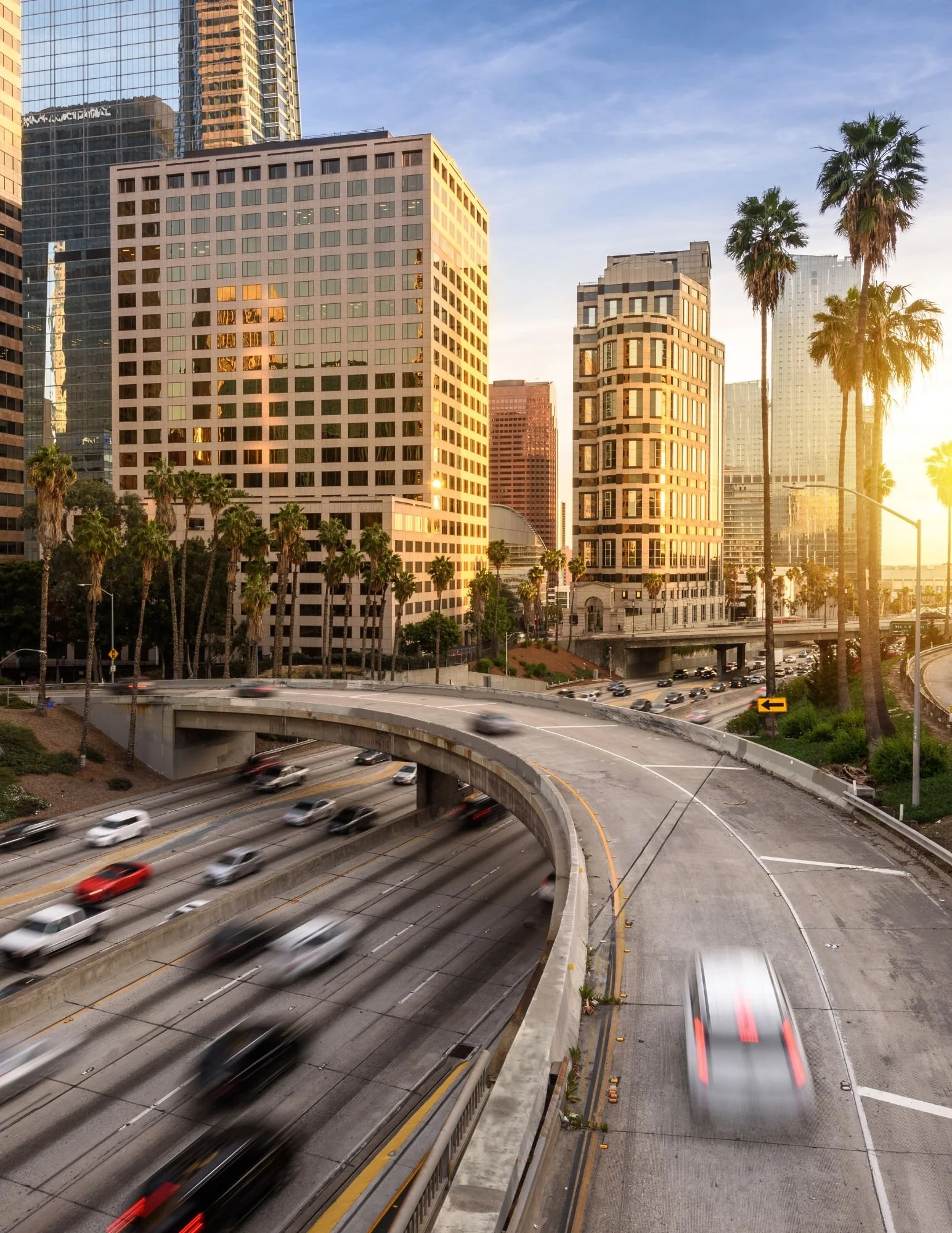 Sunset view of high-rise buildings and palm trees along a busy freeway with moving cars in Los Angeles.