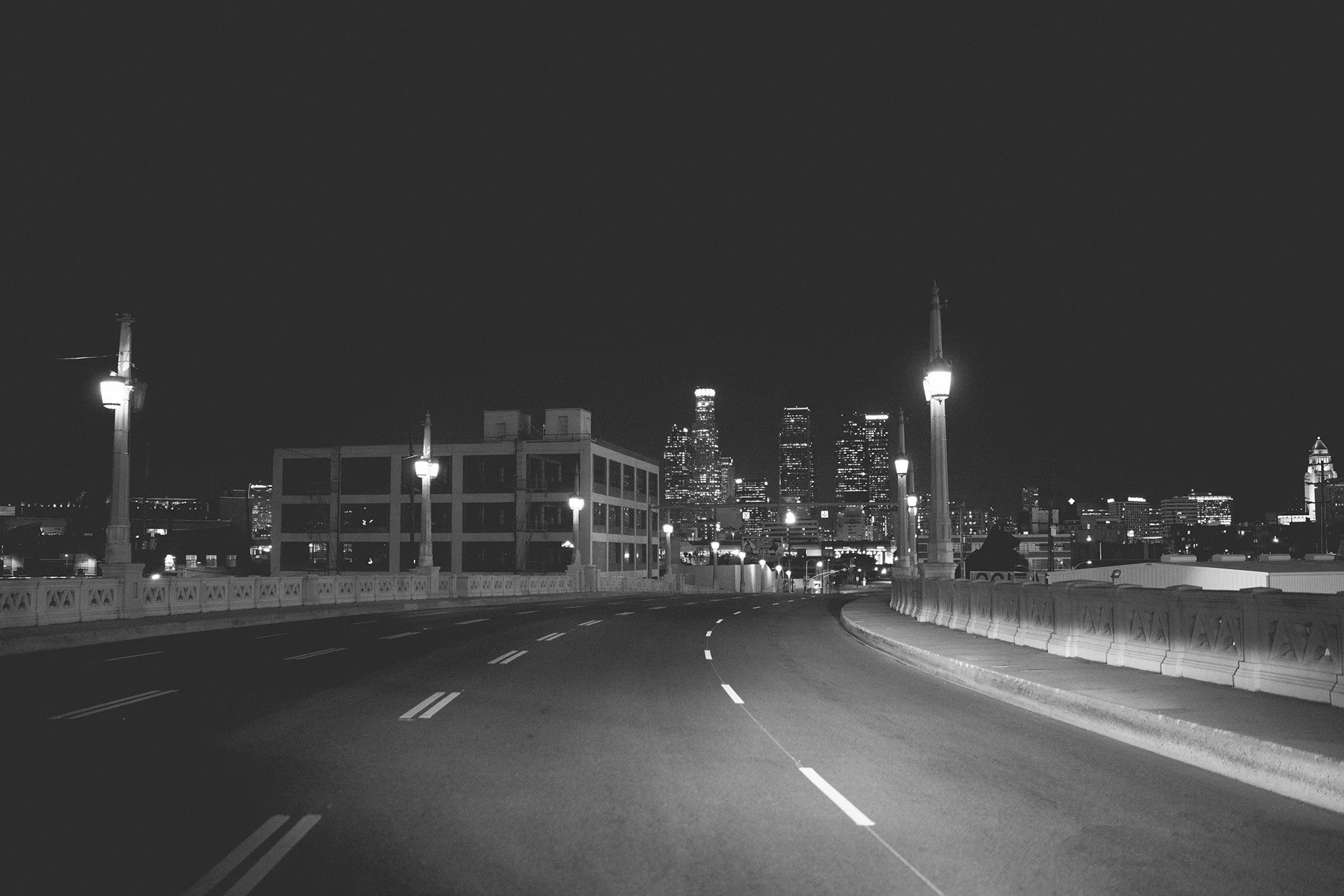 Nighttime cityscape view from a bridge with streetlights, overlooking a downtown skyline with lit skyscrapers.