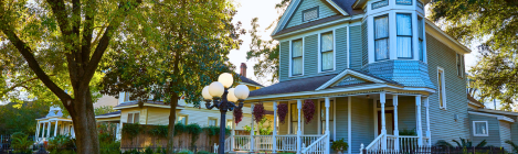 A large blue Victorian-style house with a wrap-around porch, surrounded by trees and a front yard with a lamp post.