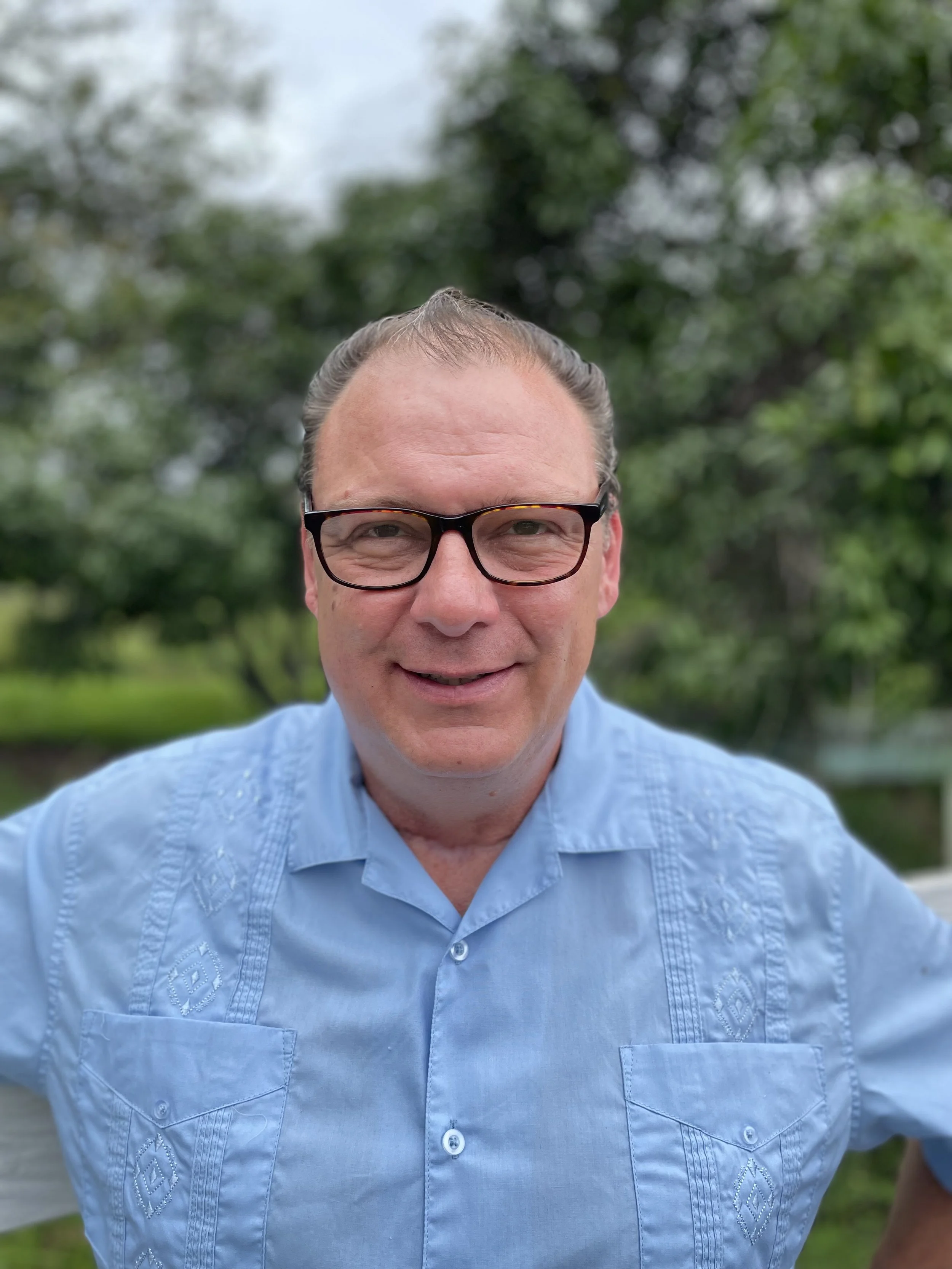 A man with light skin, glasses, and slicked-back hair is smiling outdoors, with trees and greenery in the background.