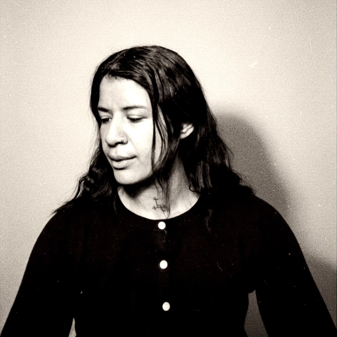 A black-and-white photo of a woman with long dark hair, looking downward and slightly to her left, wearing a dark sweater with buttons and a cross necklace, against a plain light wall background.