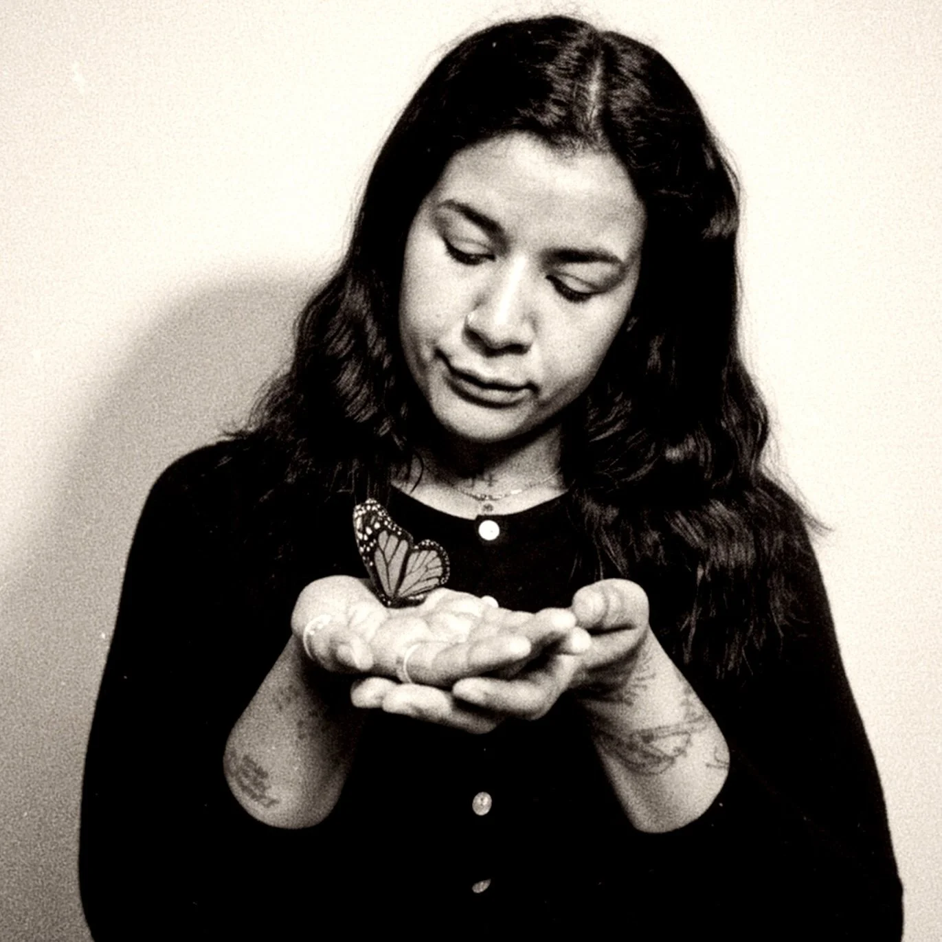A woman with long wavy hair looking down at her hands, which hold a butterfly. She is wearing a dark top with buttons, necklaces, and visible tattoos on her arms. The photo is in black and white.
