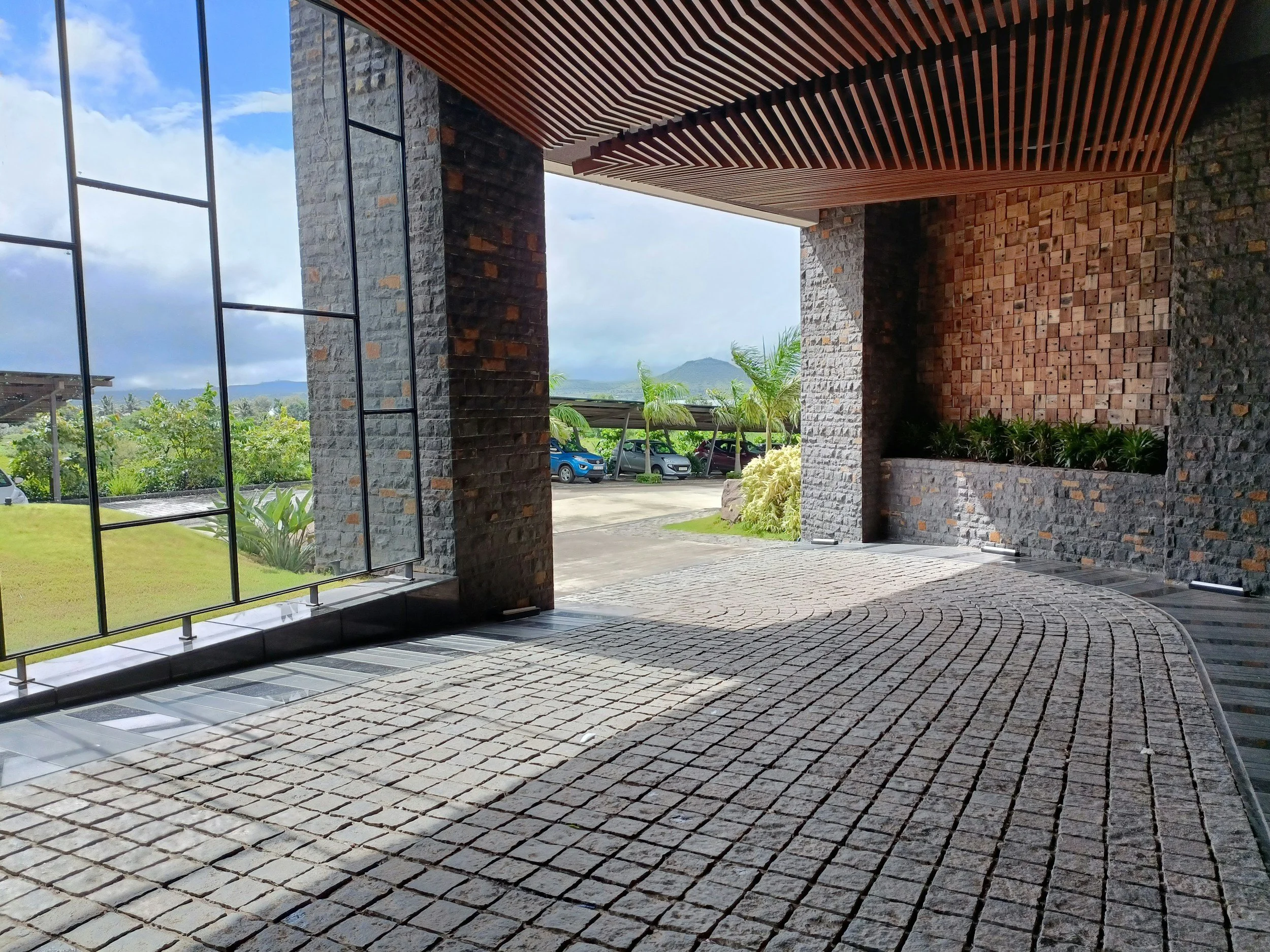 A modern building featuring large glass windows, stone and brick walls, and a wooden ceiling design. Outside, there are parked cars and greenery with a mountain and cloudy sky in the background.