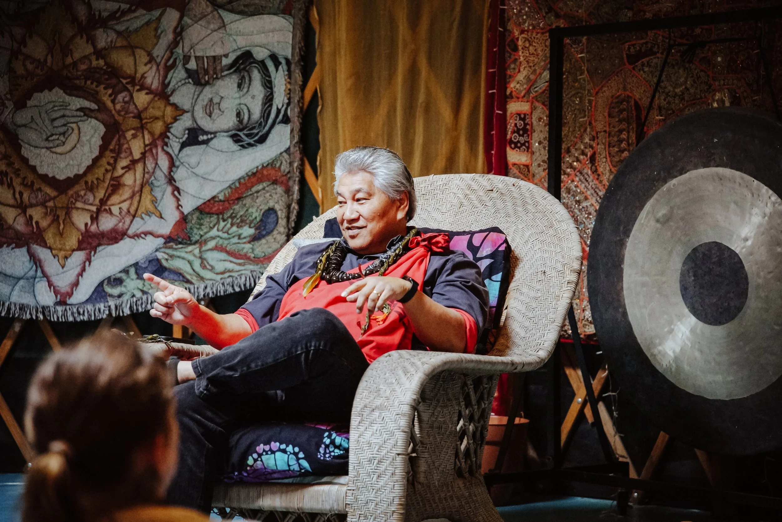 A person with gray hair, dressed in a dark and red outfit with large jewelry, relaxing in a woven chair, with colorful textiles and gongs in the background.