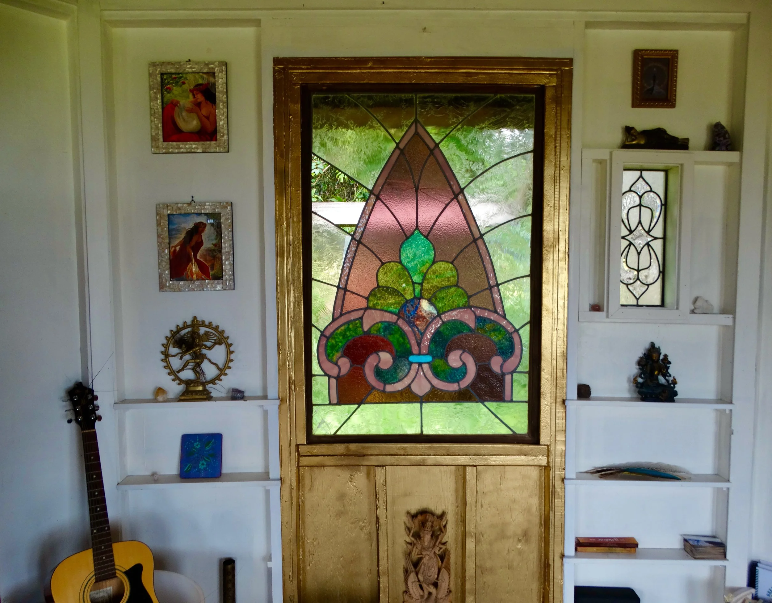 Interior of a room with a stained glass window in the center, flanked by white built-in shelves with various decorations and artwork, and a guitar leaning against the wall on the left side.
