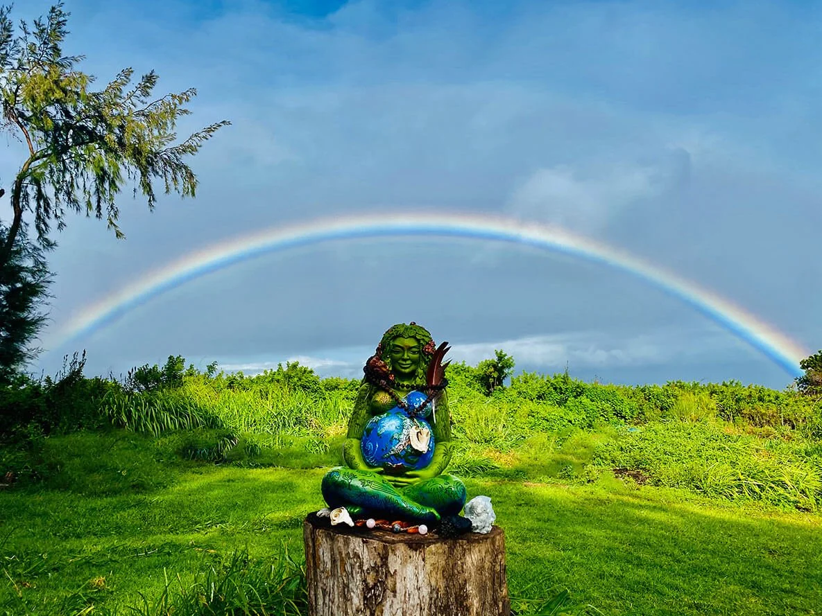 A green statue of a girl with long hair, sitting cross-legged on a tree stump, holding a blue and green globe, with a rainbow arching in the cloudy sky behind her and lush green grass and trees surrounding the scene.