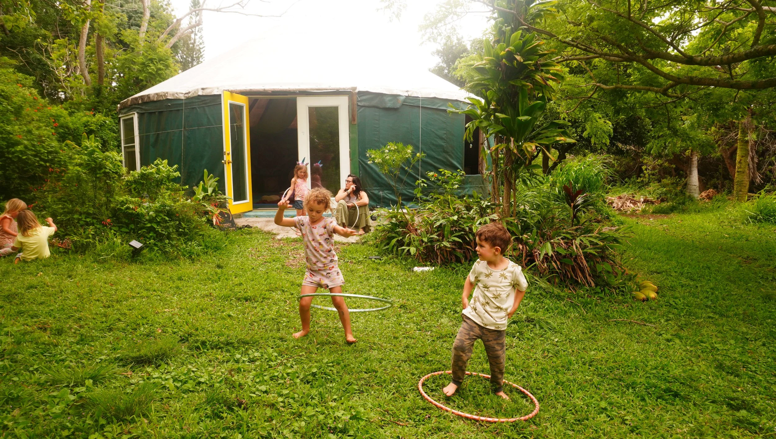 Children playing outside on a grassy area in front of a green tent or yurt surrounded by trees and lush greenery.