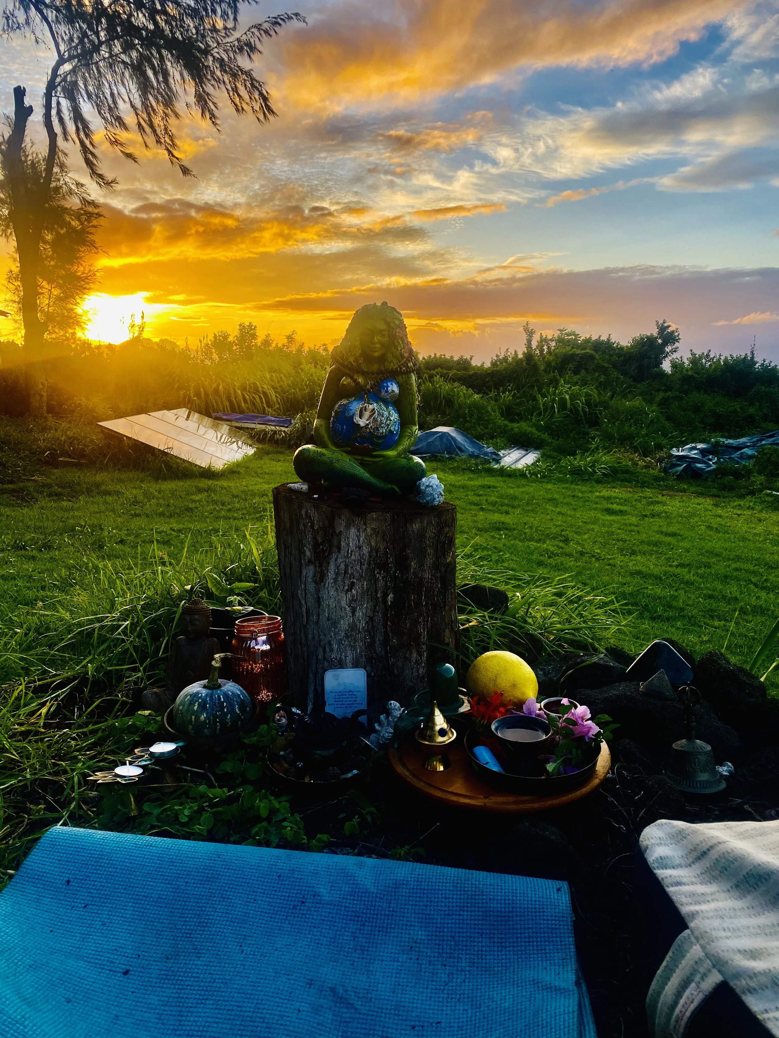 Sunset over a grassy field with a small shrine featuring a seated figure holding a globe, surrounded by candles, flowers, and other offerings, with trees and a cloudy sky in the background.