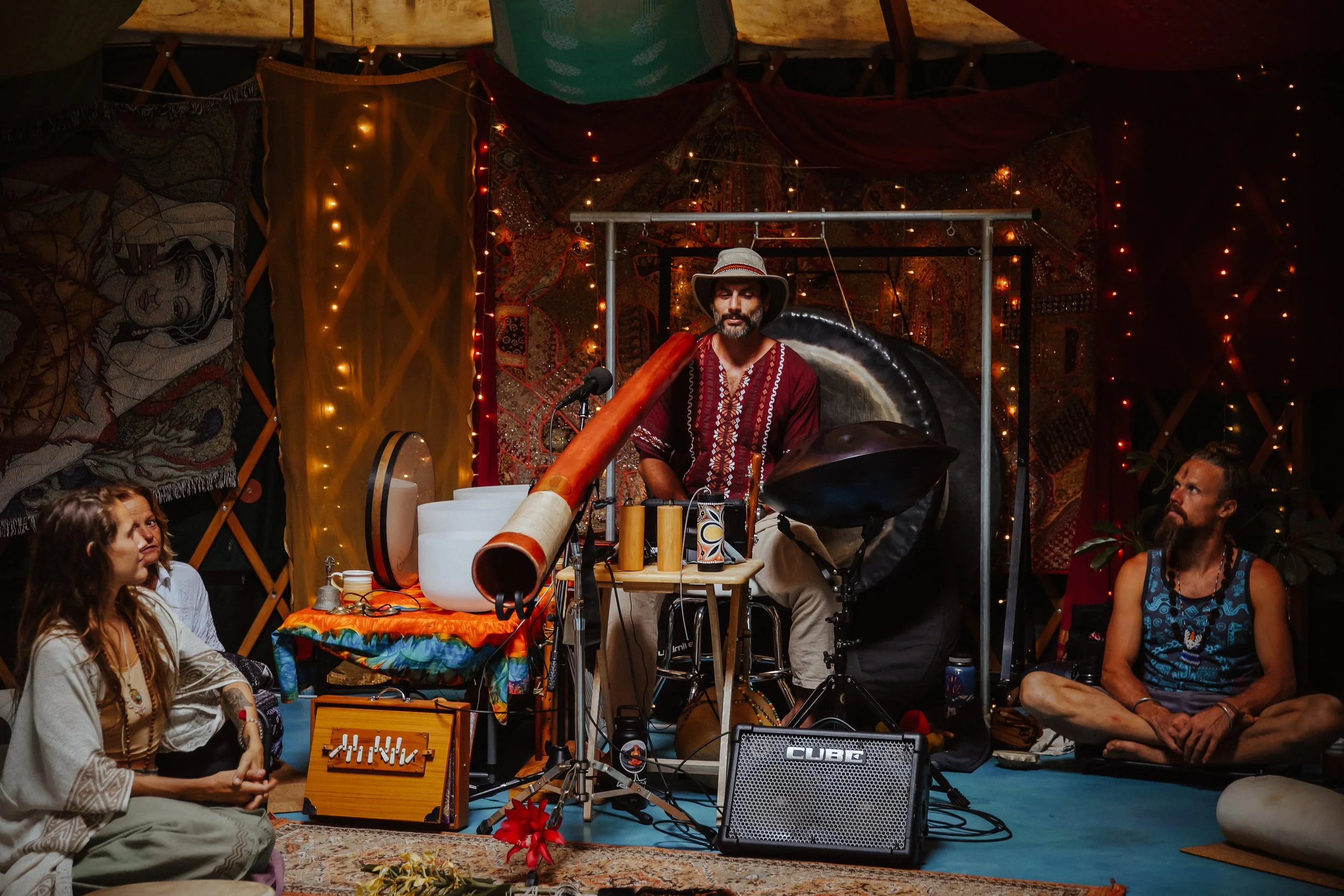 A man in a hat and patterned shirt playing a didgeridoo during a performance with people seated around him, surrounded by colorful textiles and string lights.