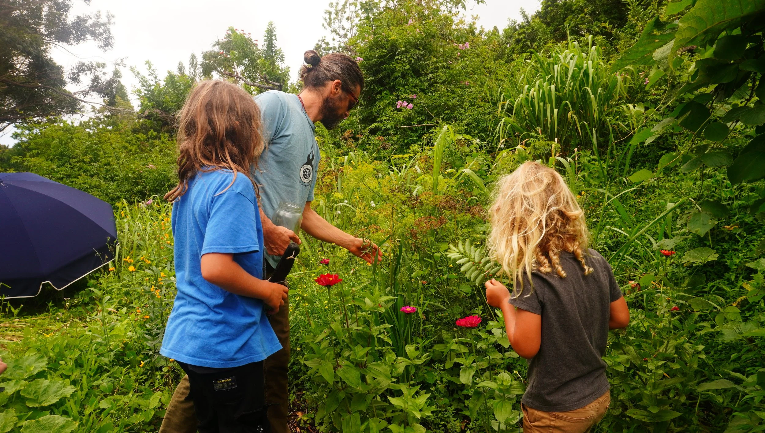 Three children and one adult girl with long, curly blond hair and dark clothing, and a man with glasses, beard, and hair tied back, in a lush green garden with pink flowers. The man is showing the girls a plant while the children look on.