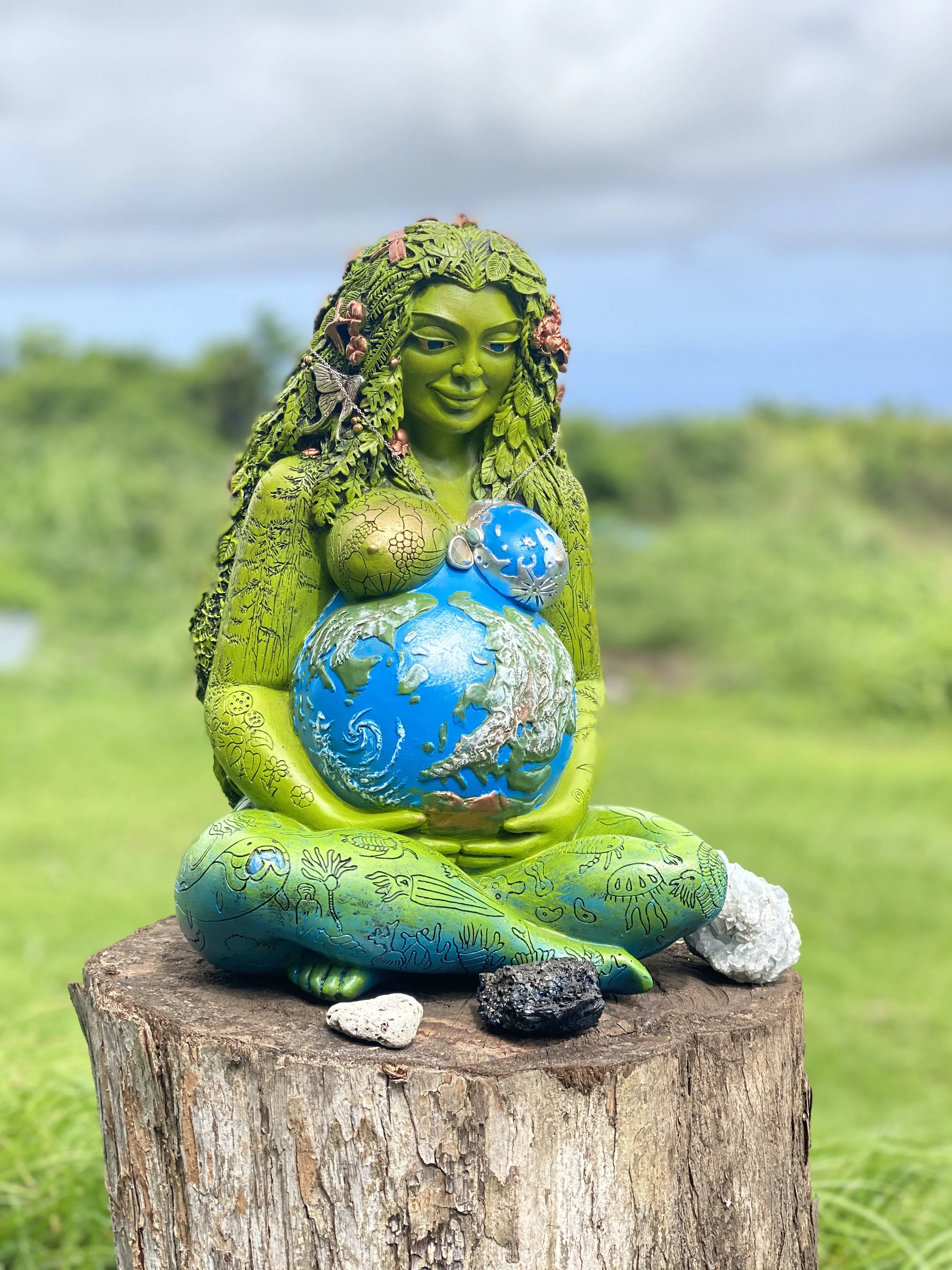 A sculpture of a woman with green skin, long hair made of leaves and adorned with insects and flowers, sitting cross-legged on a tree stump, holding a blue and green representation of the Earth, with a cloudy sky and greenery in the background.