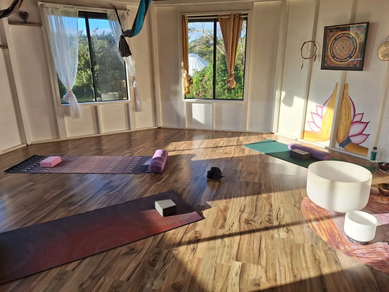 A yoga or meditation studio with wooden floors, several yoga mats and props, and large windows showing greenery outside. The room has a peaceful, natural ambiance with decorative art on the walls.