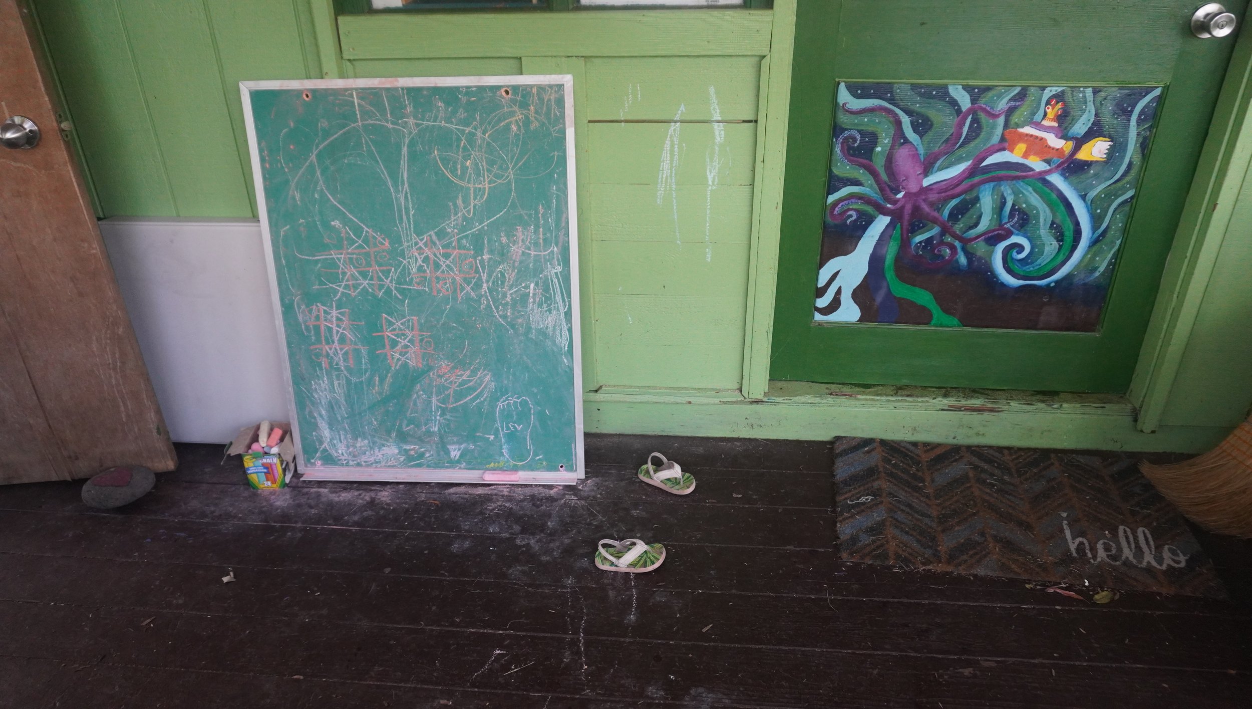 A green wooden wall with a chalkboard, a colorful painting of an octopus, a pair of sandals, some chalk, and a rock on a weathered wooden floor.