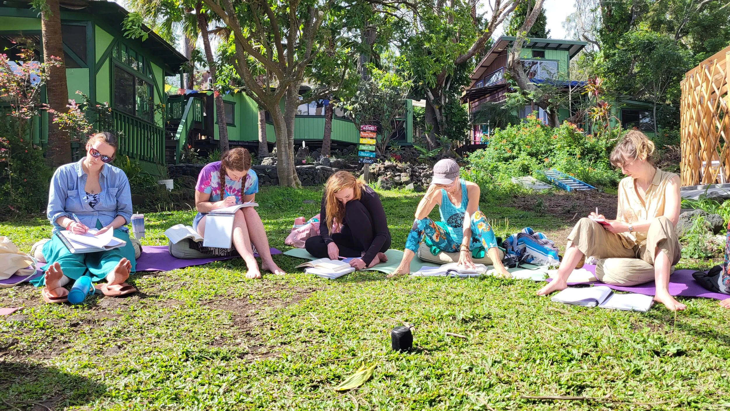 A group of six women sitting on mats and cushions on grass in a garden, reading and writing in notebooks, surrounded by trees and plants, with step houses in the background.