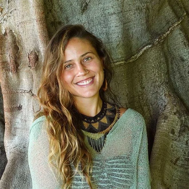 A smiling woman with long wavy hair, wearing a light green sweater and a beaded black and gold necklace, standing against a large tree trunk.