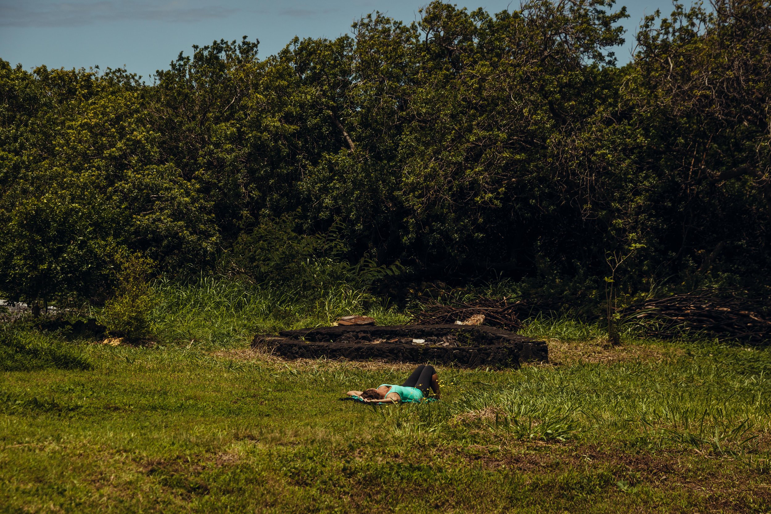 A person lying on a grassy field, facing downward, wearing a teal shirt and black pants, with a forested area in the background and a clear blue sky above.