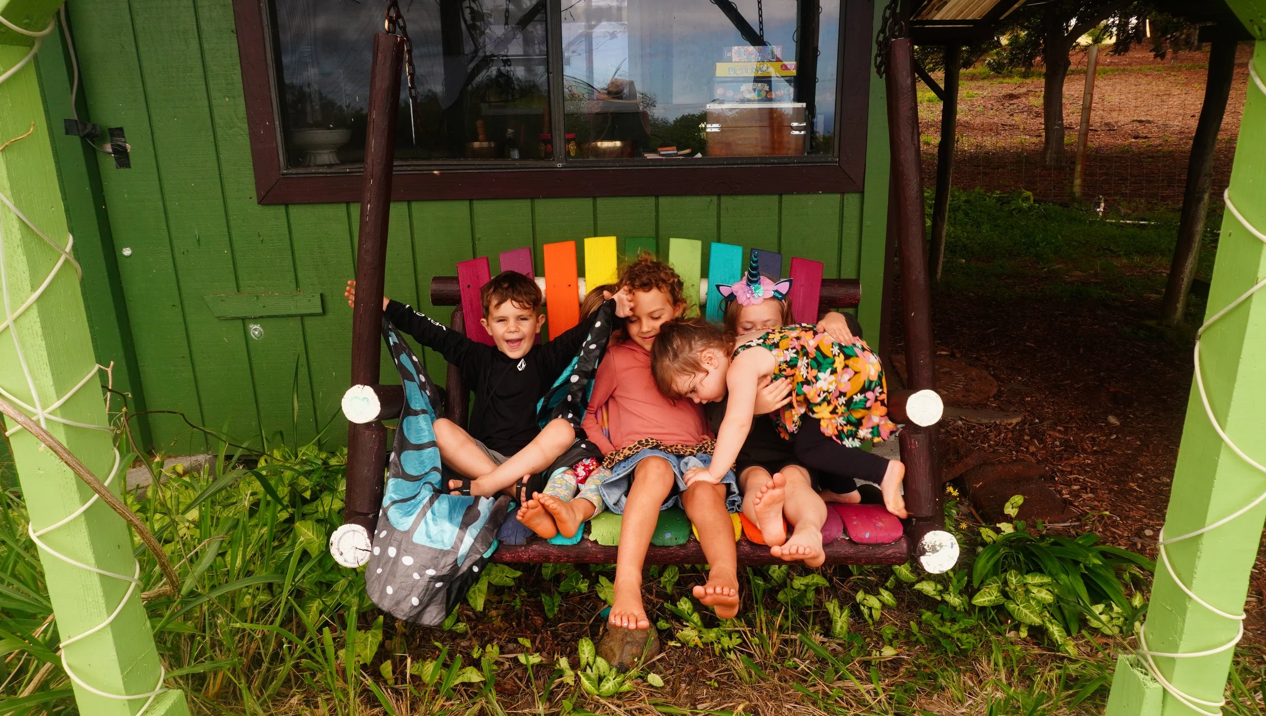 Four children sitting on a wooden swing with a colorful backrest, hugging and laughing together. The swing is outdoors in a garden setting with green plants and trees, and a building with a window in the background.