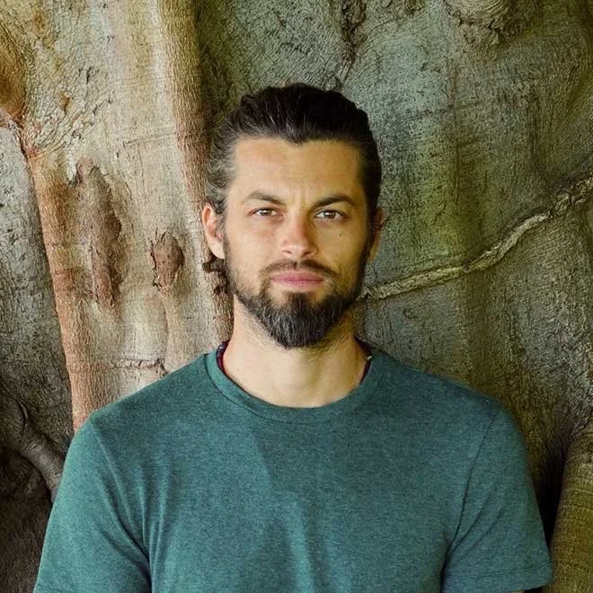 A man with a beard and long hair tied back, standing outdoors against a tree trunk.