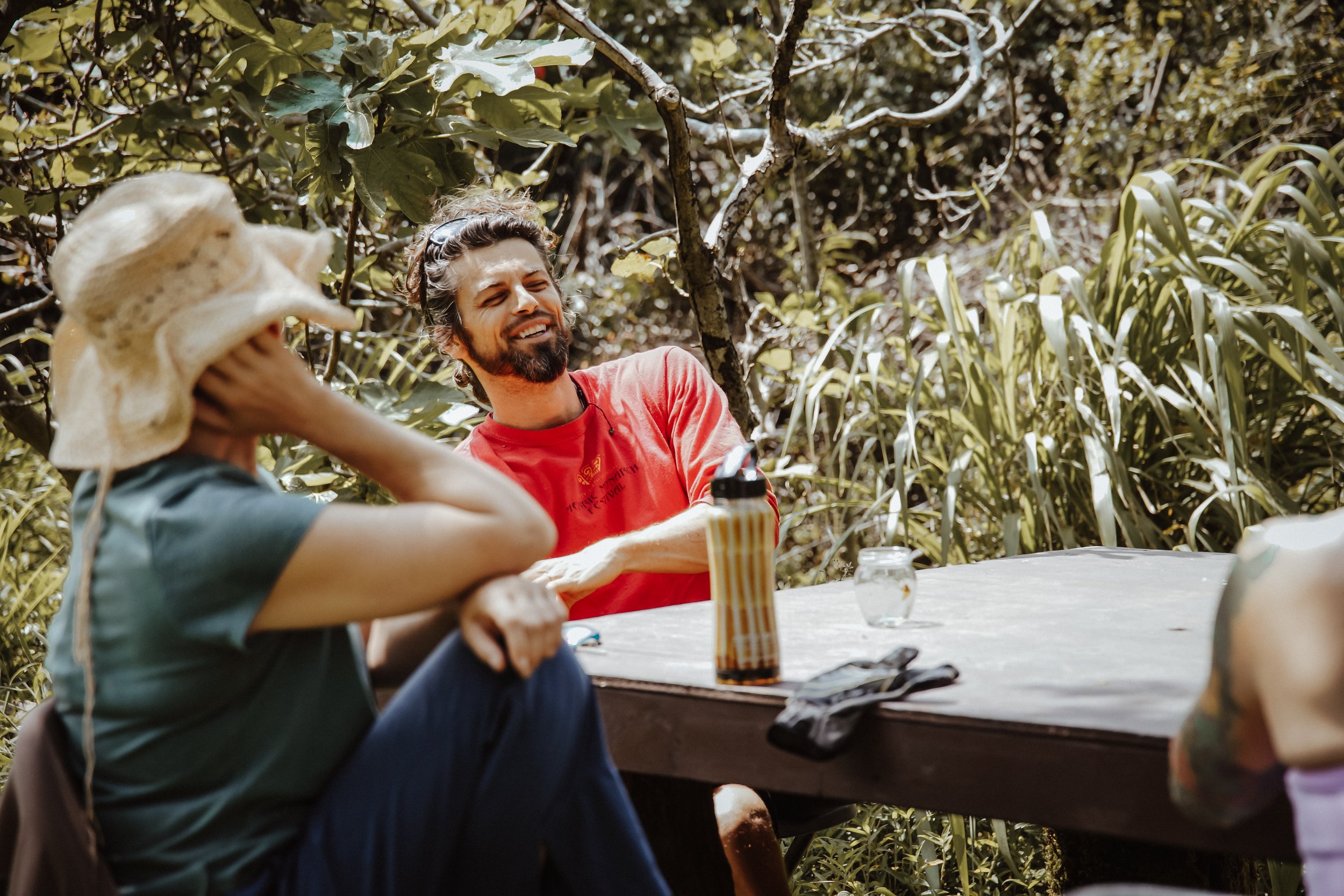 Two people sitting at a picnic table outdoors, one woman wearing a straw hat and the man smiling in a red shirt, surrounded by greenery and trees.