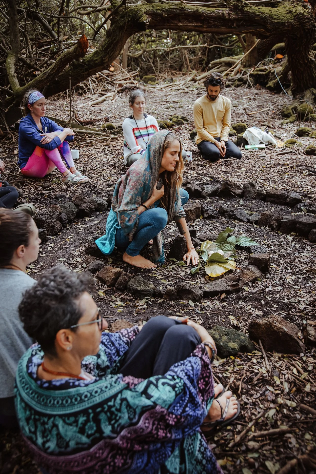 Group of people sitting on the forest ground, some with eyes closed, gathered around a central area with leaves, stones, and a small arrangement of natural objects, possibly engaging in a meditation or ritual.