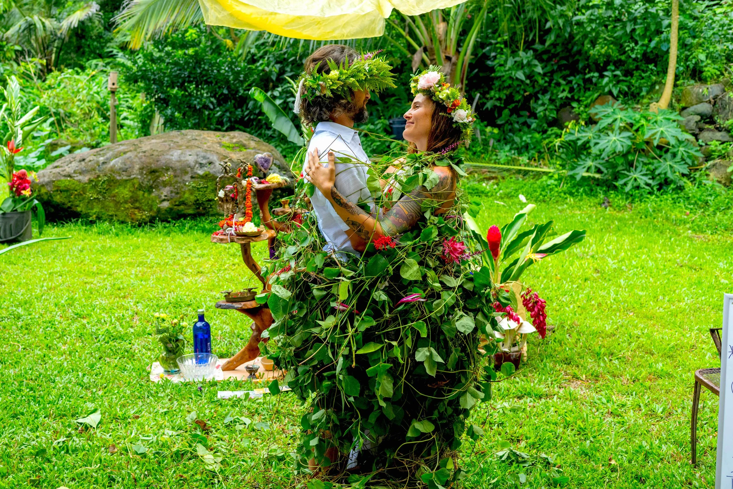 A couple dressed in ceremonial attire, adorned with flower crowns and surrounded by greenery, sharing a joyful moment outdoors.
