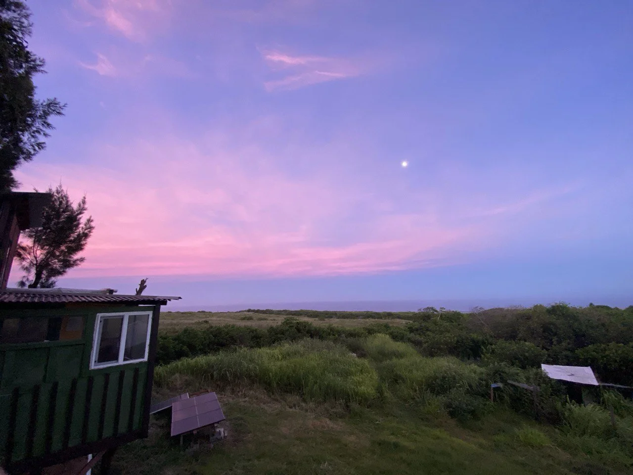 A panoramic view of a rural landscape at dusk with a colorful pink and purple sky, a visible moon, green bushes and trees, and small structures including a green shed and a solar panel.