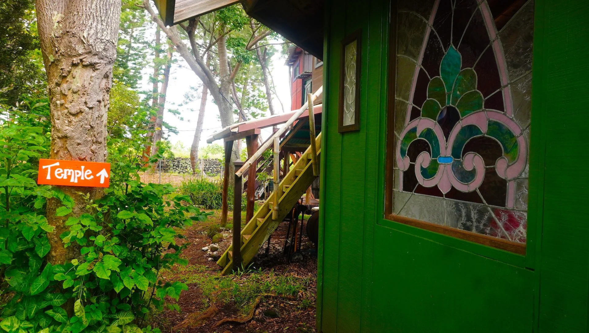 A green building with a stained glass window, surrounded by trees and plants. A wooden staircase leads up to the building, and there is an orange sign on a tree that reads 'Temple' with an arrow pointing upward.
