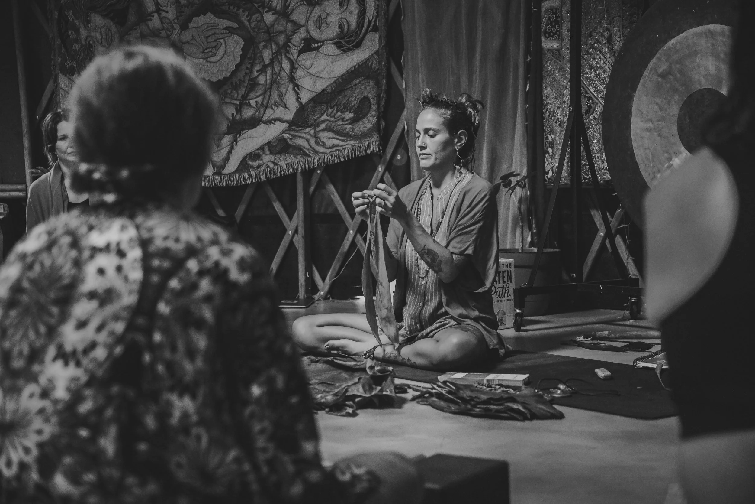 A woman with tattoos and styled hair, sitting cross-legged on a mat, appears to be teaching or demonstrating something to a small group in an art or yoga studio, with artwork and textiles hanging on the walls.