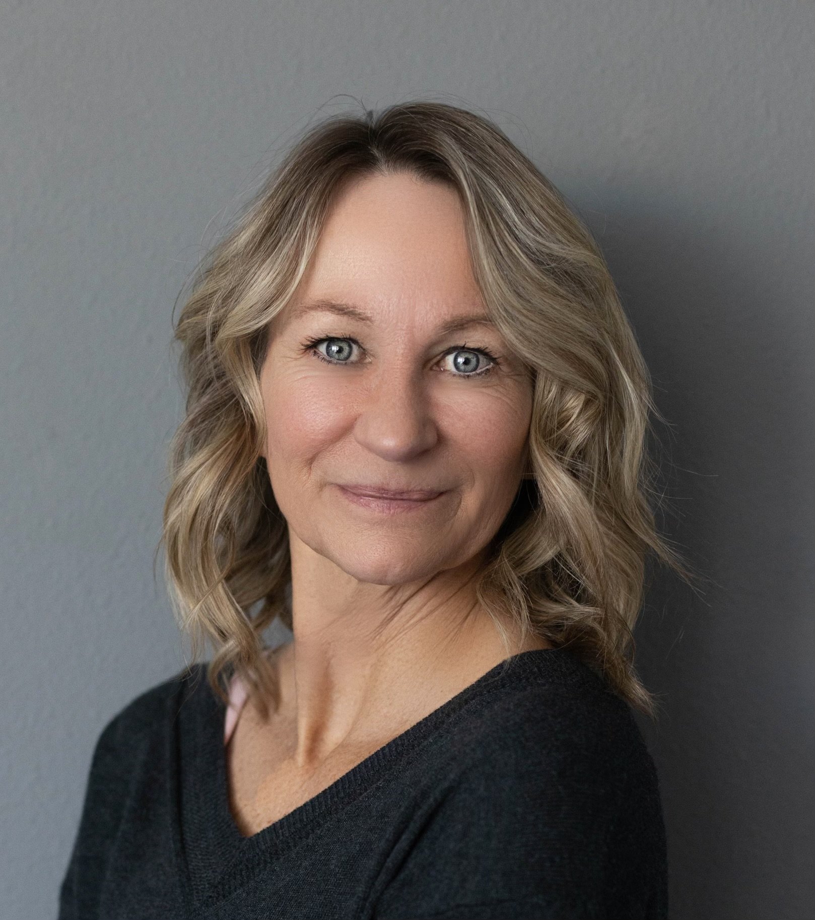 Jody, a woman with blonde, wavy hair and blue eyes, wearing a black top, stands against a gray wall.