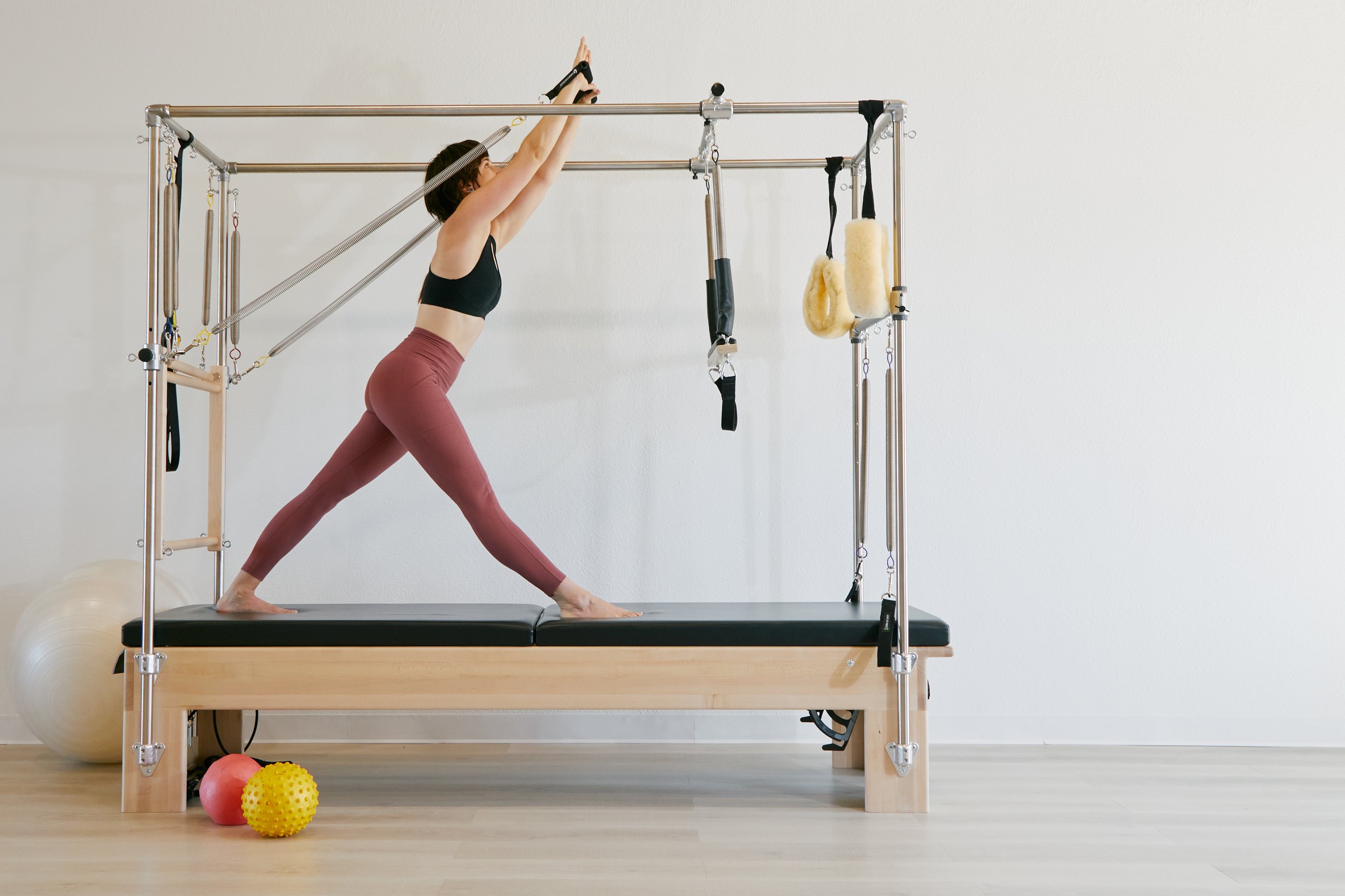 A woman in workout clothes performing a yoga pose on a Pilates reformer machine in a bright, minimalist room. There are exercise balls and foam rollers on the floor.