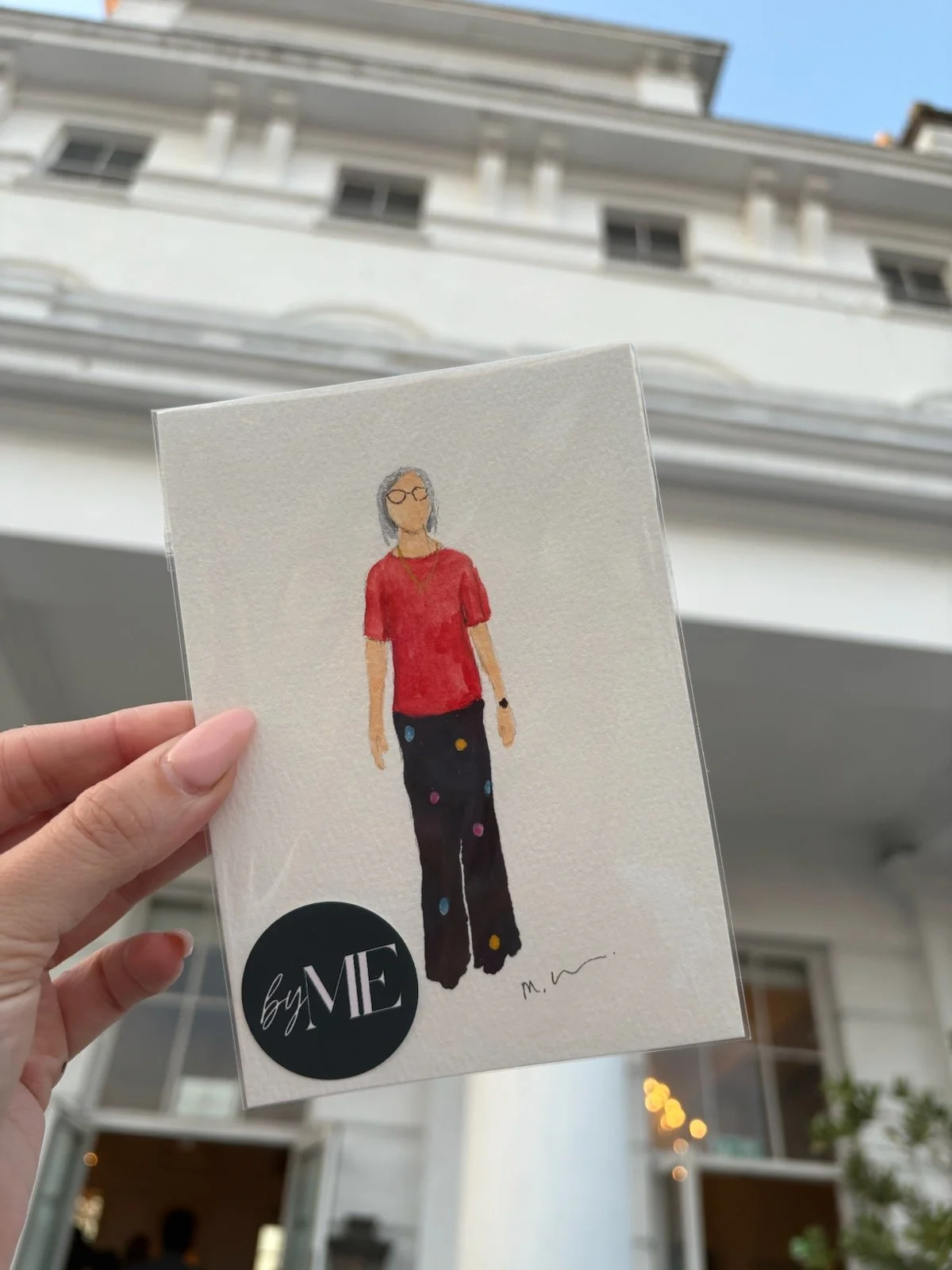 A hand holding a greeting card with a watercolor illustration of a woman wearing glasses, a red top, and black pants with colorful dots. The background shows a white building with balcony railings and windows against a blue sky.
