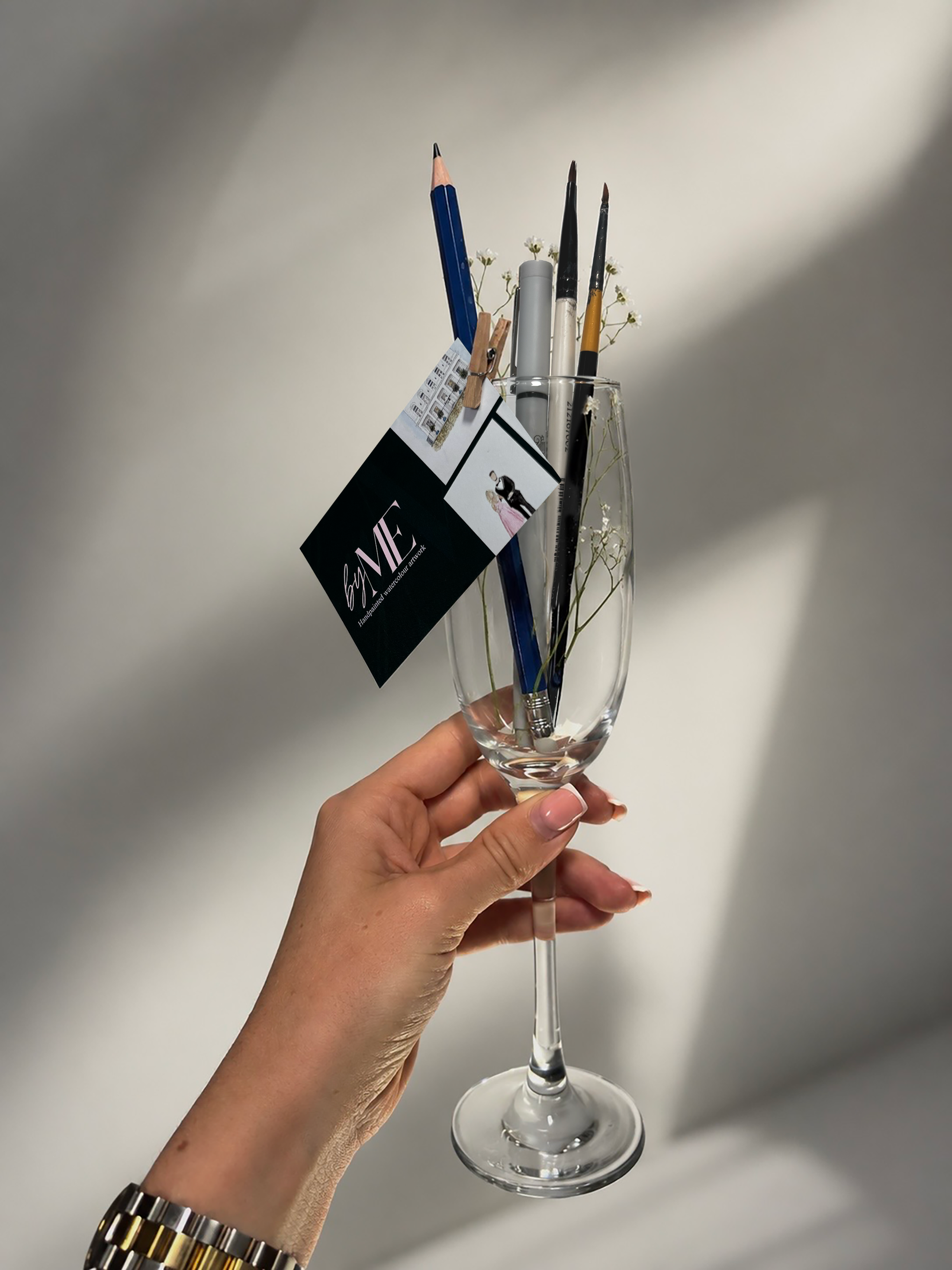 A hand holding a champagne flute filled with small white gypsophilia, two paint brushes, a pencil and a pen. A dark green business card with the word 'by ME' is attached on it with a peg.
