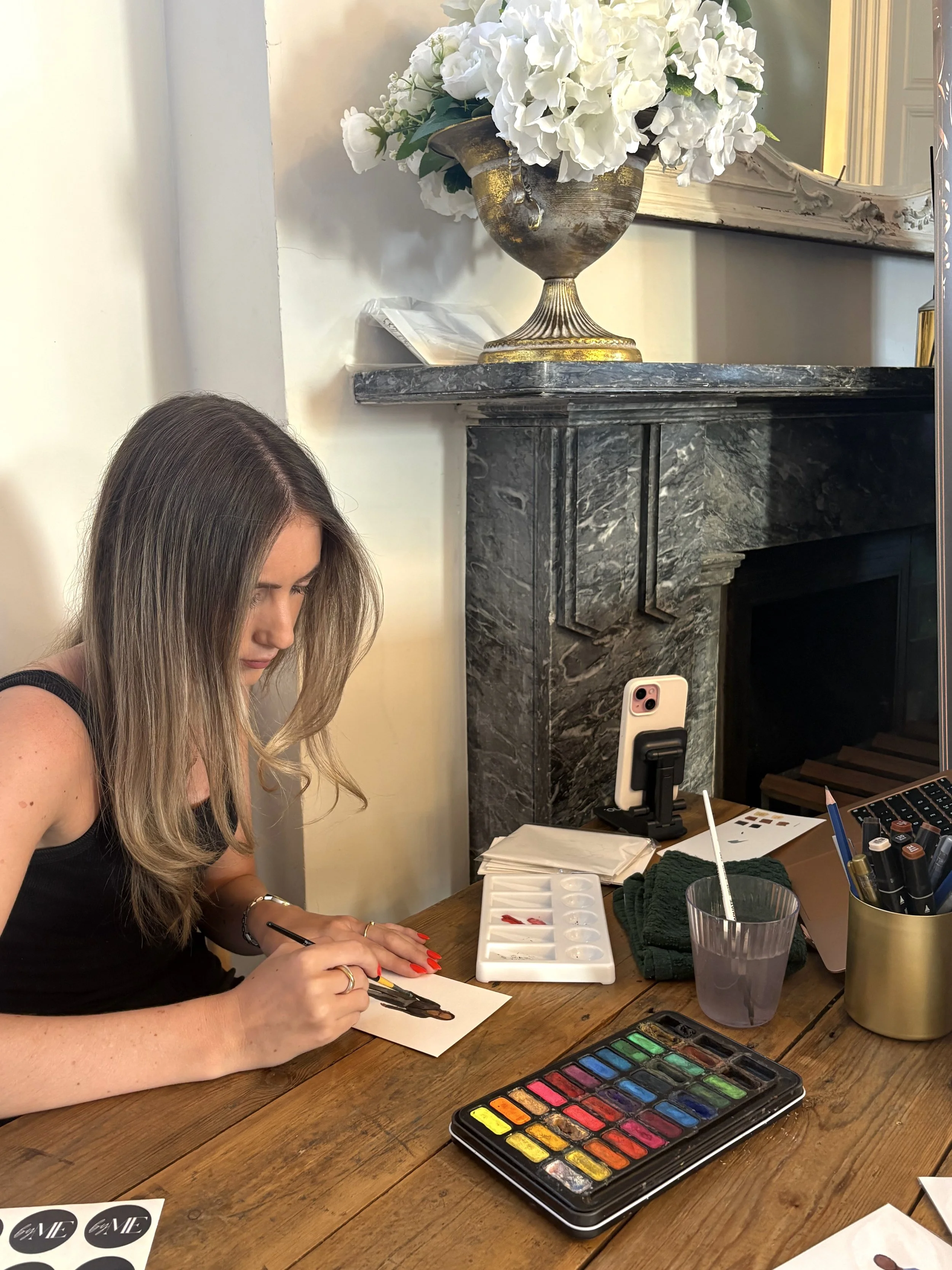 A woman with long hair and a black top is painting a picture using watercolors at a wooden table. Her left hand is holding the paper steady while her right hand uses a brush. The table also has a watercolor palette, a cup of water, a dark green cloth, and various art supplies. In the background, there is a marble fireplace with a decorative mirror and a large golden vase filled with white hydrangea flowers.