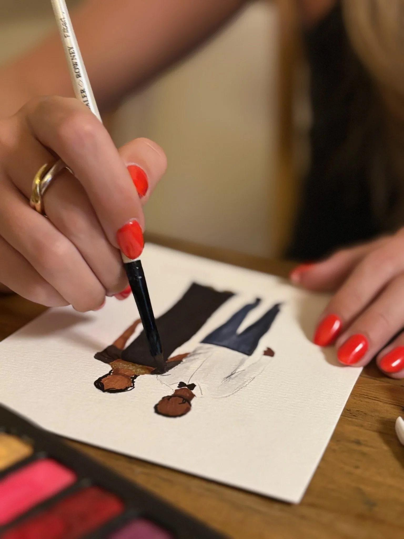 A person with red painted nails is using a black paint brush to paint on a sheet of white paper. The paper has a watercolour sketch of a two people, in brown, black, and beige. The person's hand is wearing two rings, one gold and one silver.