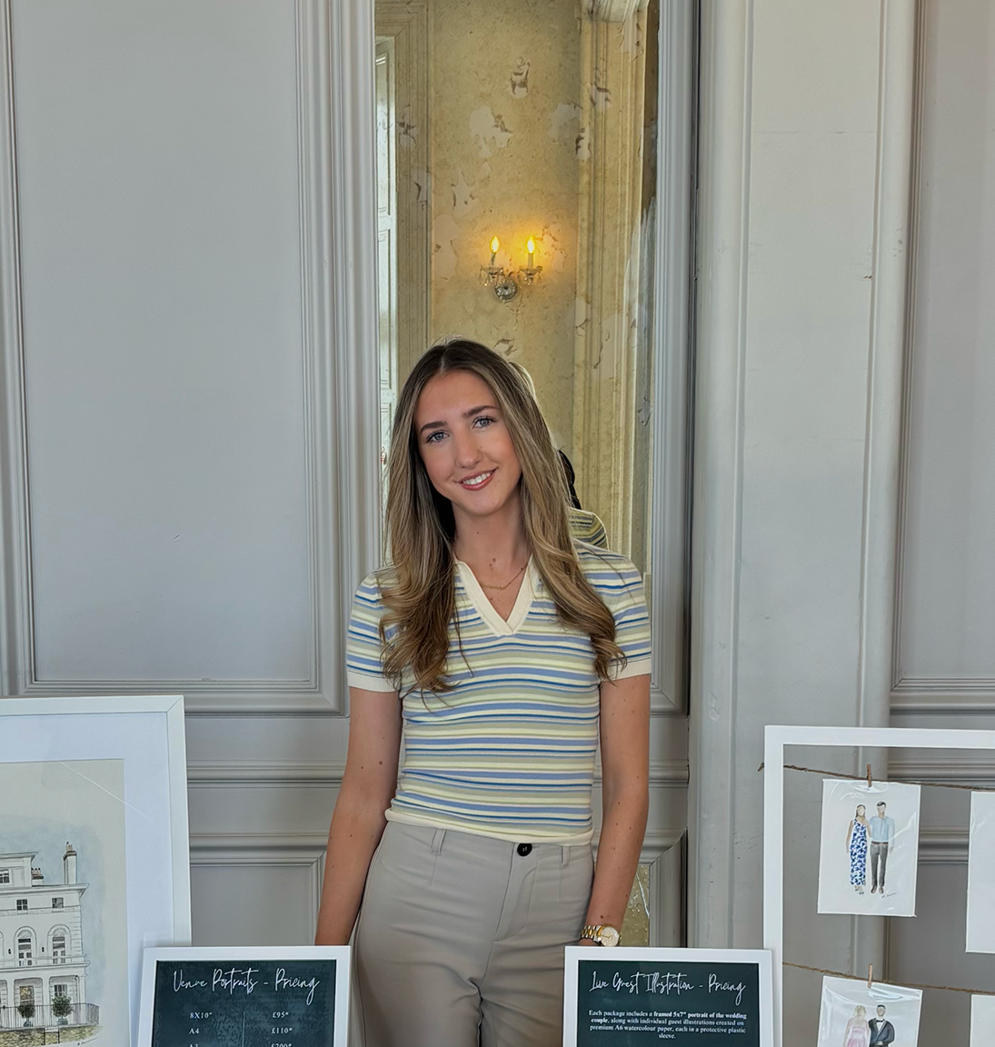 A young woman with long brown/blonde hair in a striped yellow shirt and beige pants standing behind two signboards, in a room with a decorative wall and mirror behind her, and framed drawings on display to her sides.