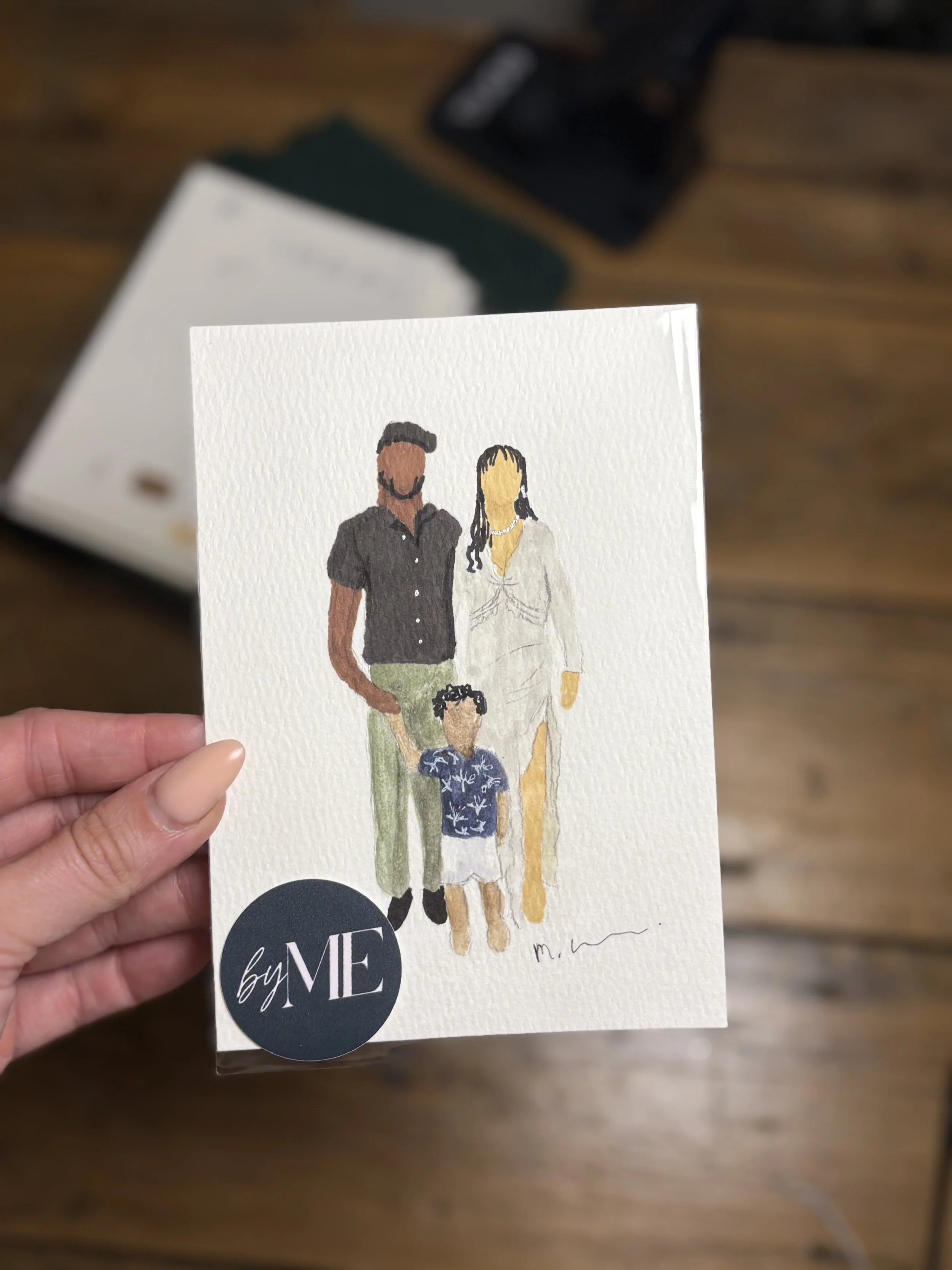 A hand holding a watercolor illustration of three people, two adults and one child, with blurred objects in the background.