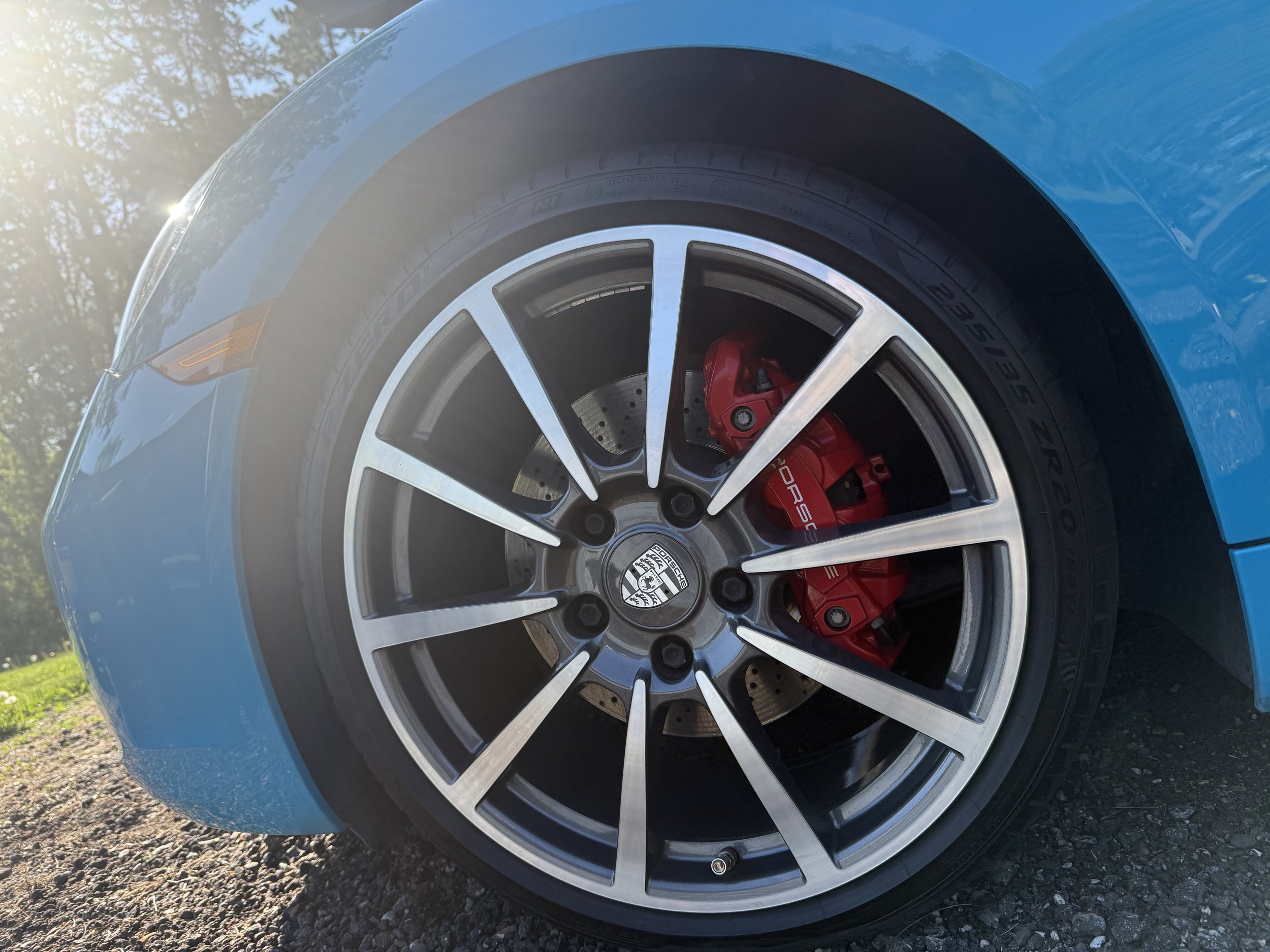 Close-up view of a blue sports car's front wheel with a silver alloy rim, red brake caliper, and ventilated brake disc, on a gravel surface with trees and sky in the background.