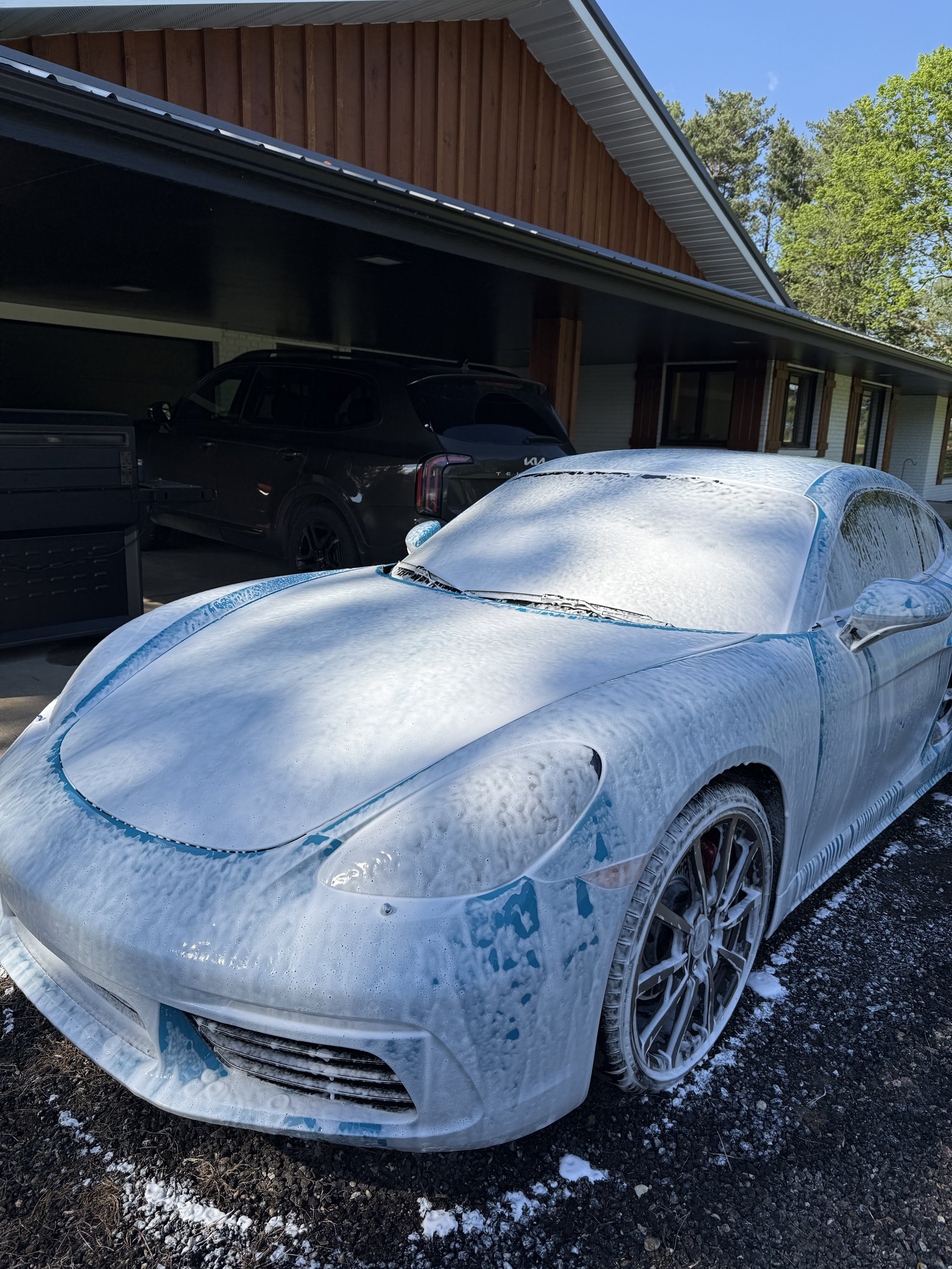 A white sports car covered in snow parked in front of a house with a covered porch. A black SUV is parked behind it, and some snow is on the ground.