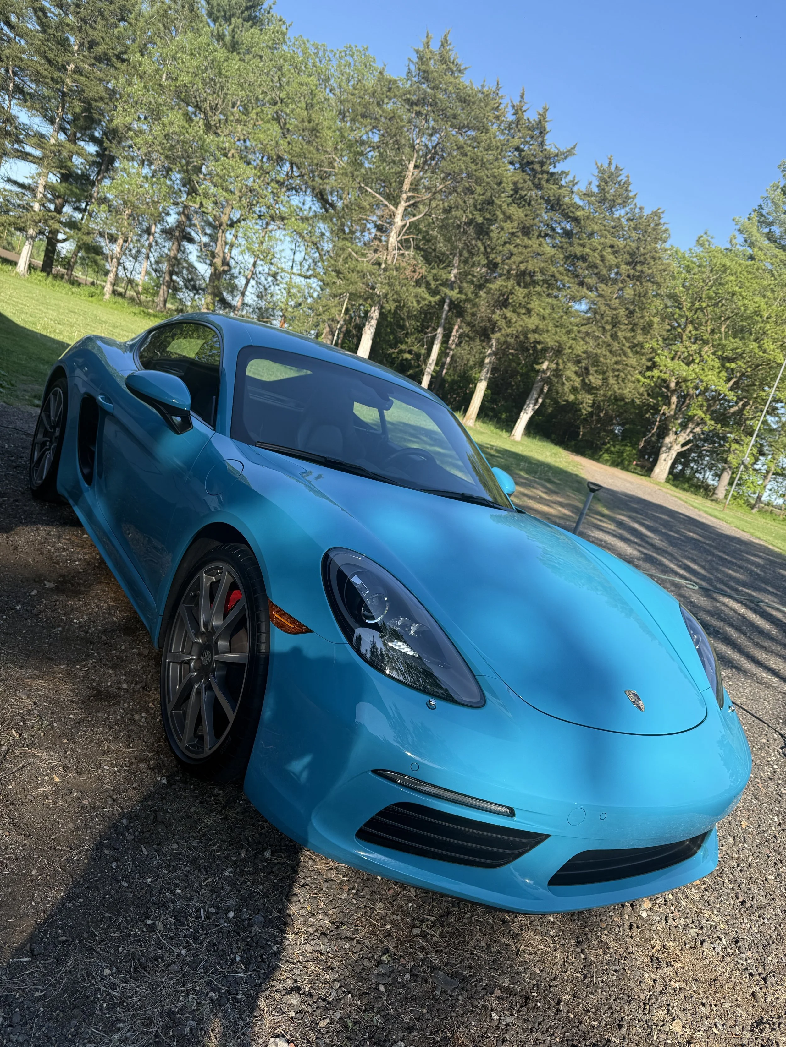 A bright blue sports car parked on a gravel surface with a wooded area and blue sky in the background.