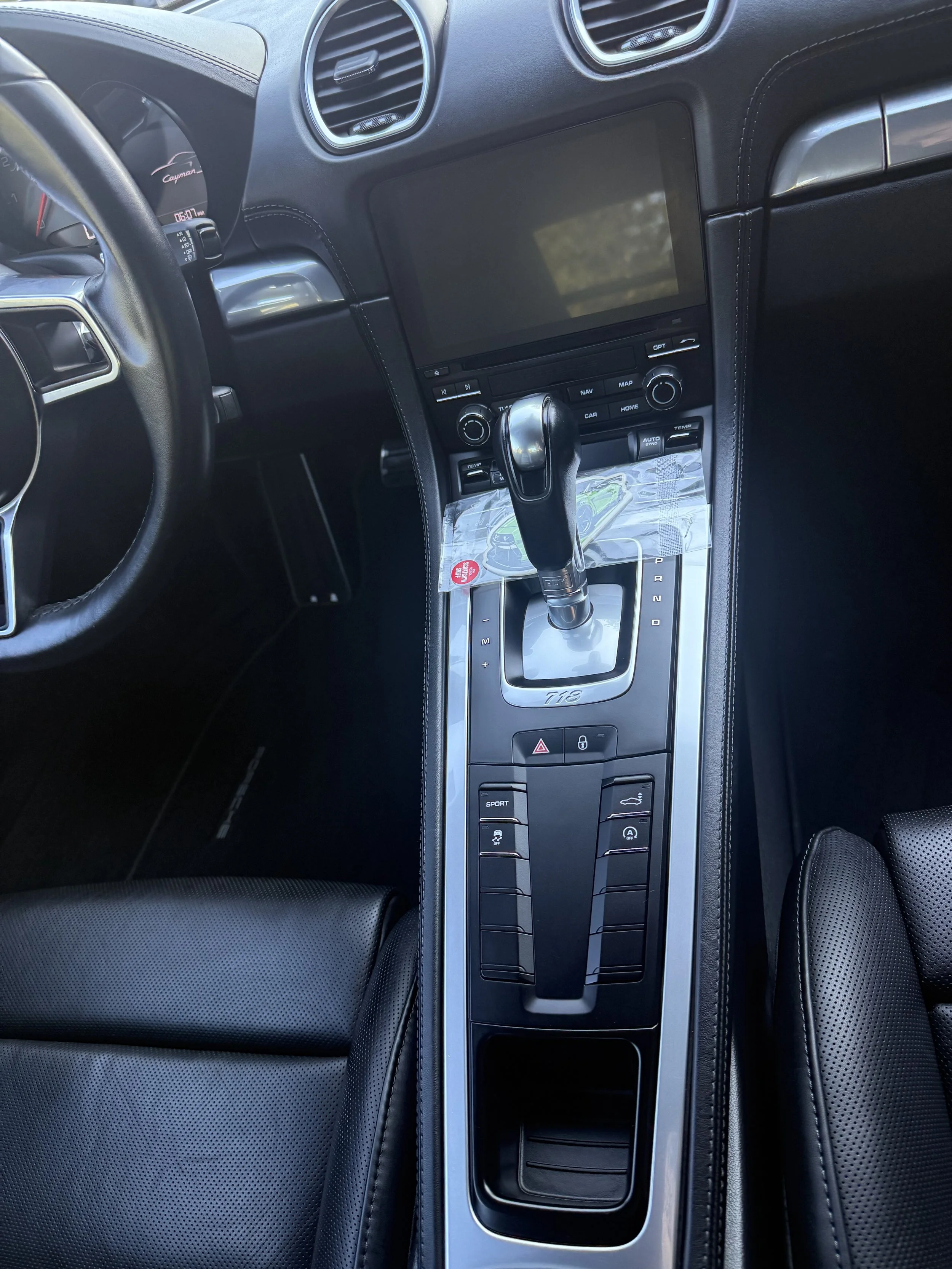 The interior of a luxury sports car, specifically a Porsche 718, showing a leather-wrapped dashboard, a central touchscreen display, an automatic gear shift, and various control buttons.