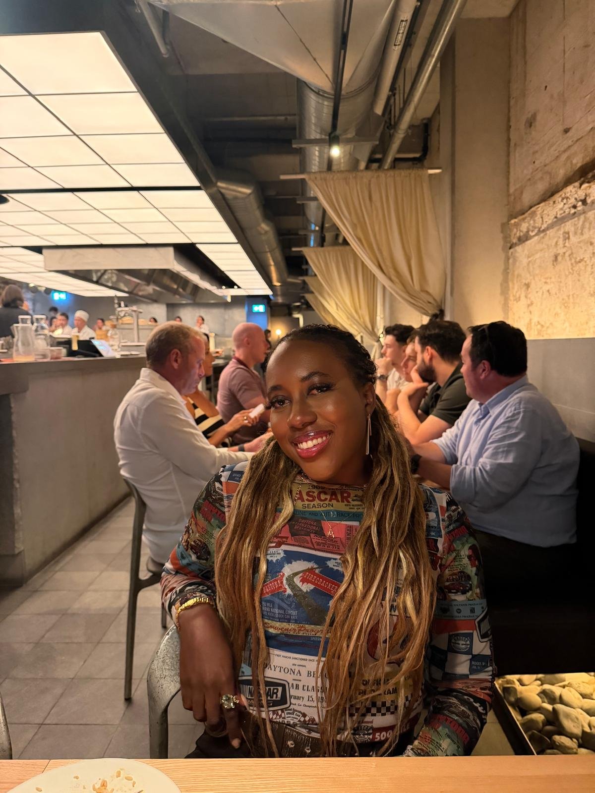 A woman with long braids, smiling, sitting in a restaurant, with people dining in the background and a bar area visible.