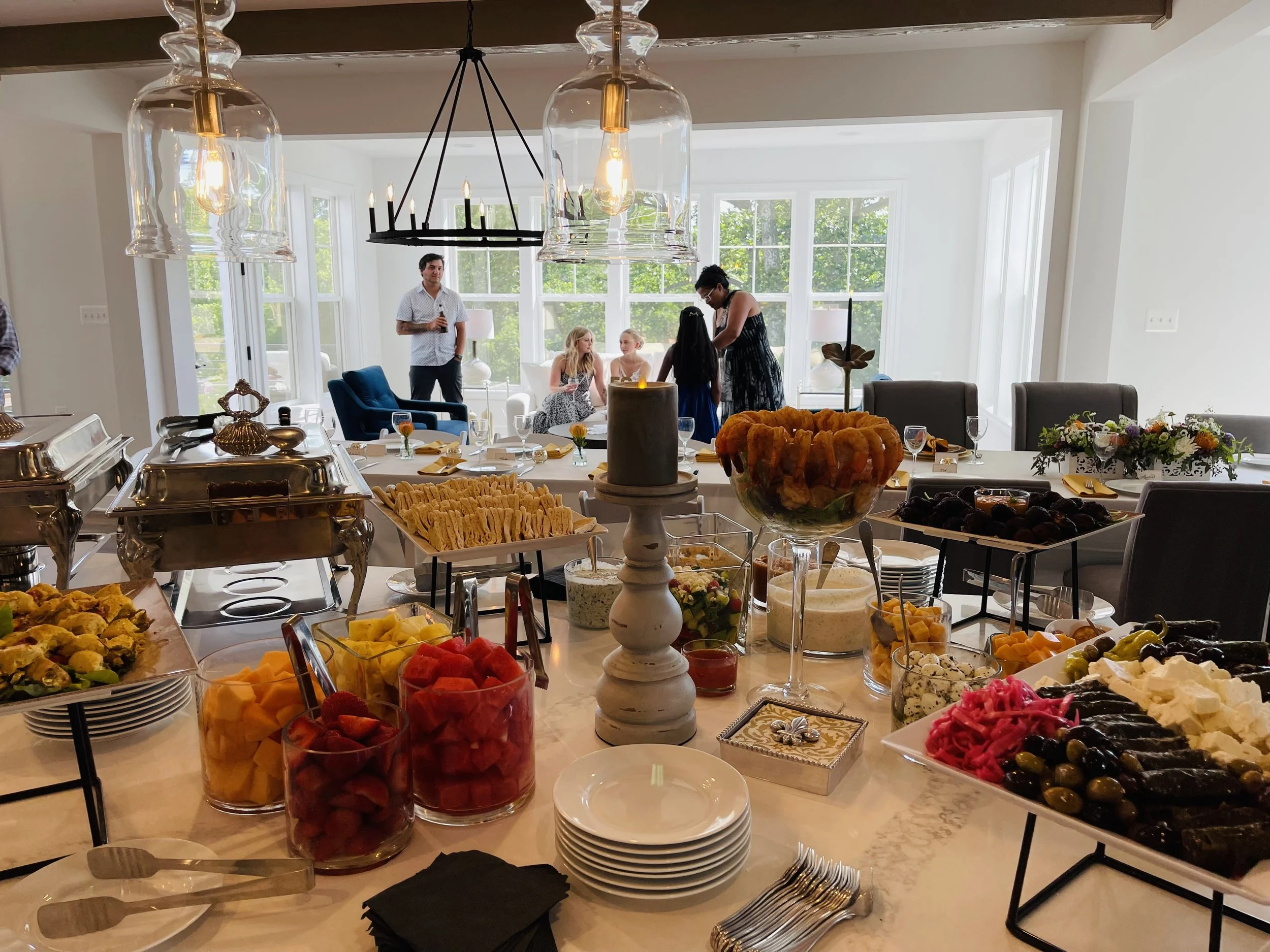 A buffet table with various fruits, cheeses, and snacks, with people standing and sitting in a bright room with large windows and modern lighting.