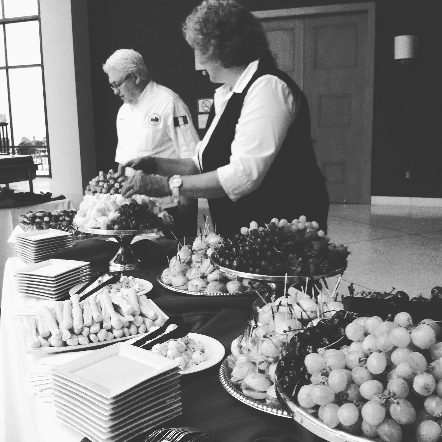Two women standing behind a table filled with various trays of fruit and appetizers in a buffet setting.