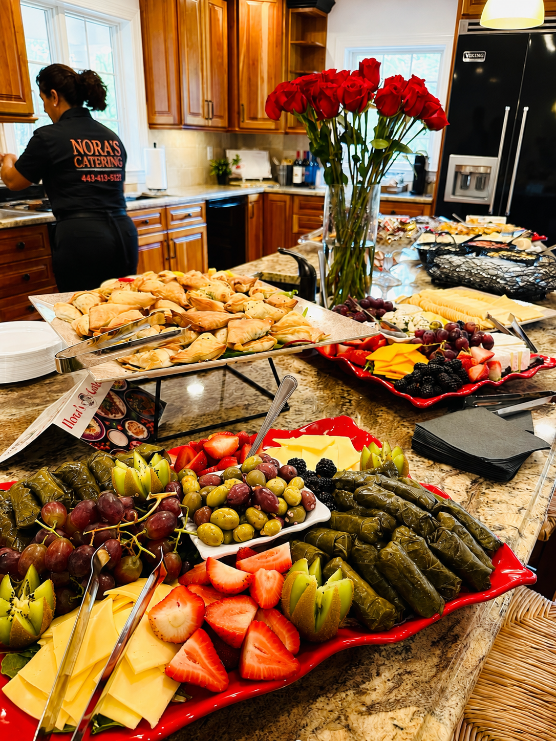 A kitchen with a catering setup featuring a large platter of fruit, including strawberries, grapes, blackberries, and sliced kiwi, along with a platter of stuffed pastries and a vase of red roses on a granite countertop.