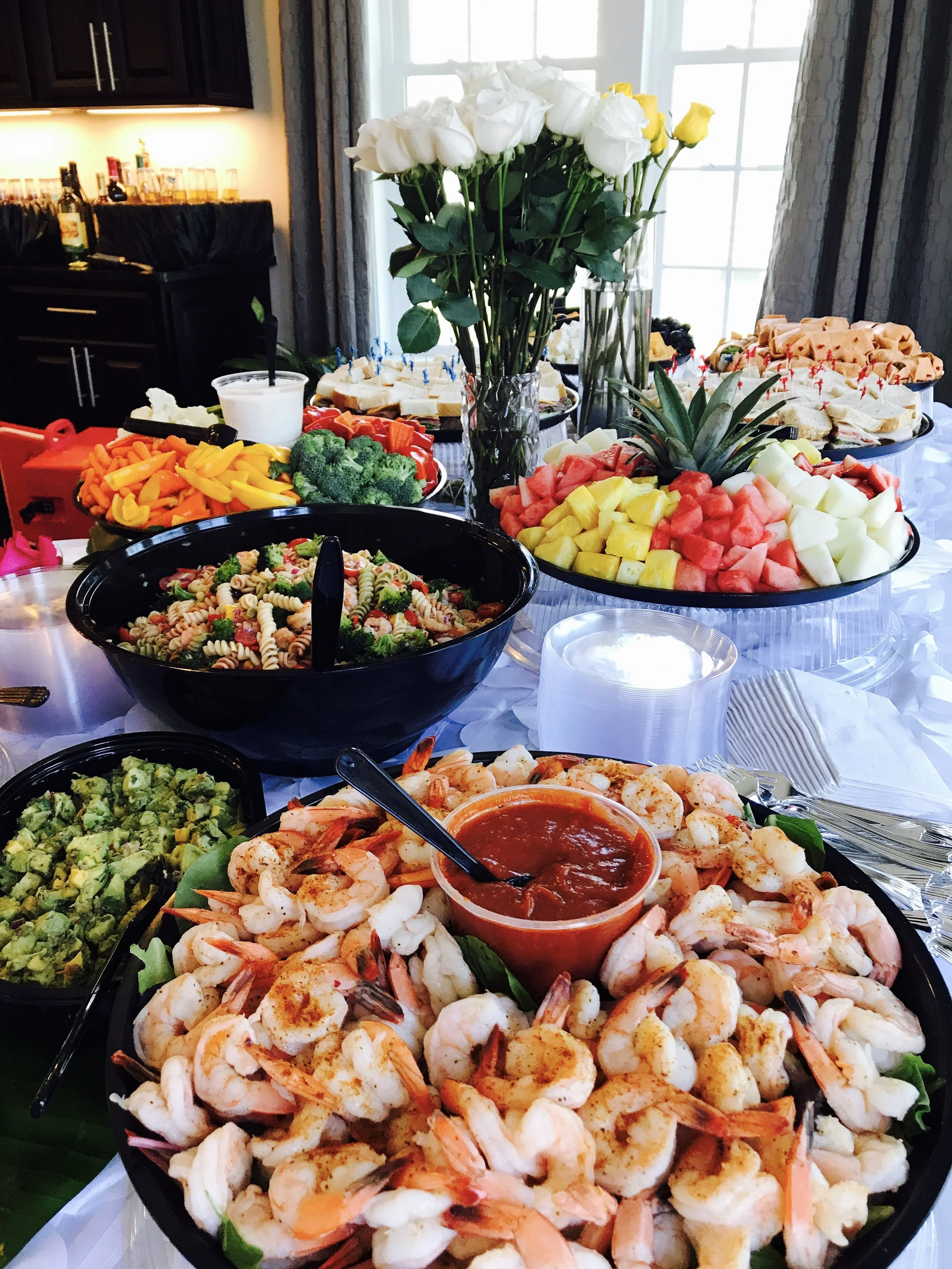 A buffet table with various foods including a bowl of cooked shrimp with a cup of cocktail sauce, fresh fruit salad with watermelon, honeydew, and pineapple, sliced vegetables, pasta salad, and sandwiches, with a vase of white roses and yellow roses in the center.