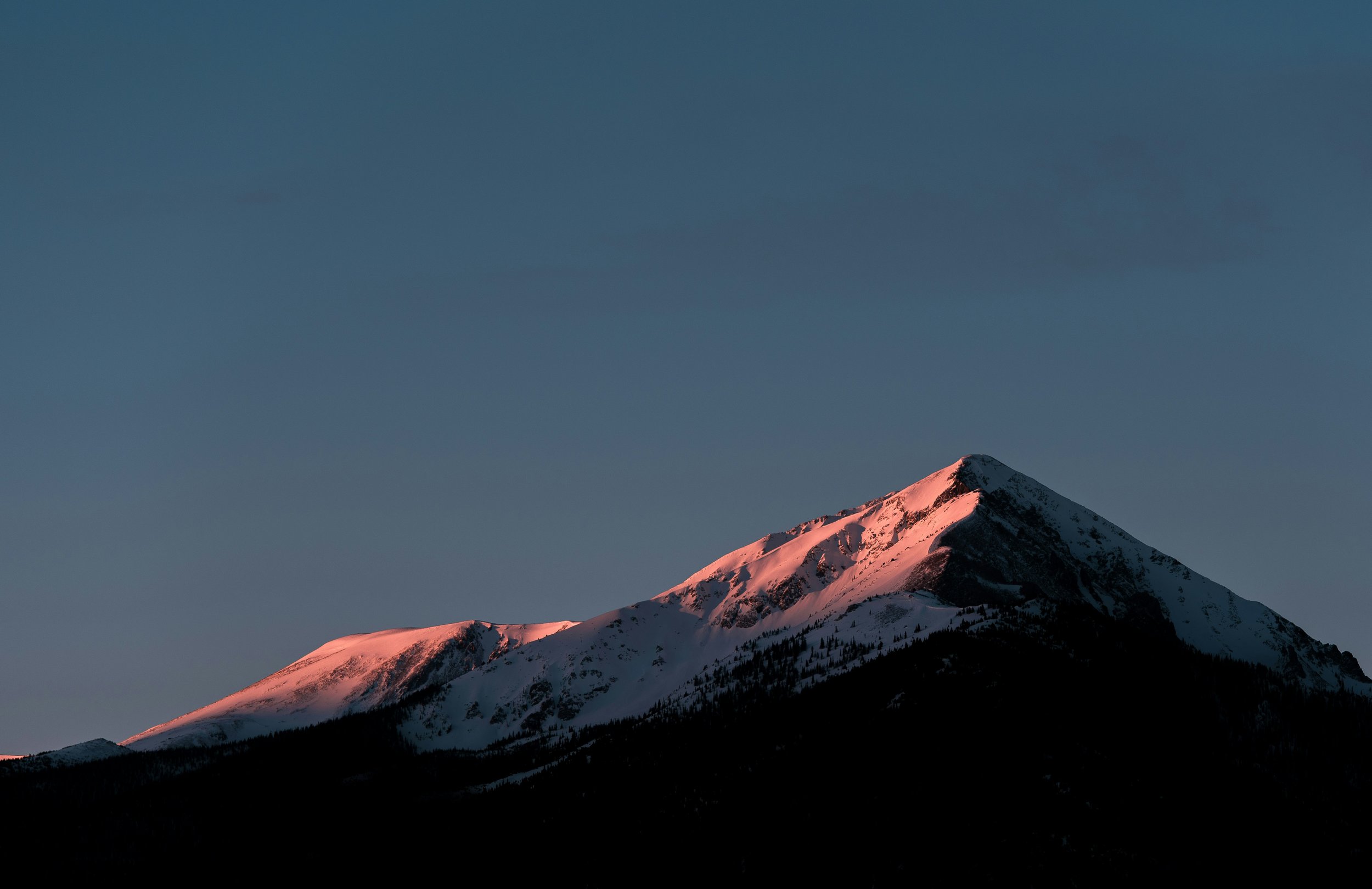 A snow-covered mountain illuminated by pink sunlight at sunset or sunrise against a clear sky.